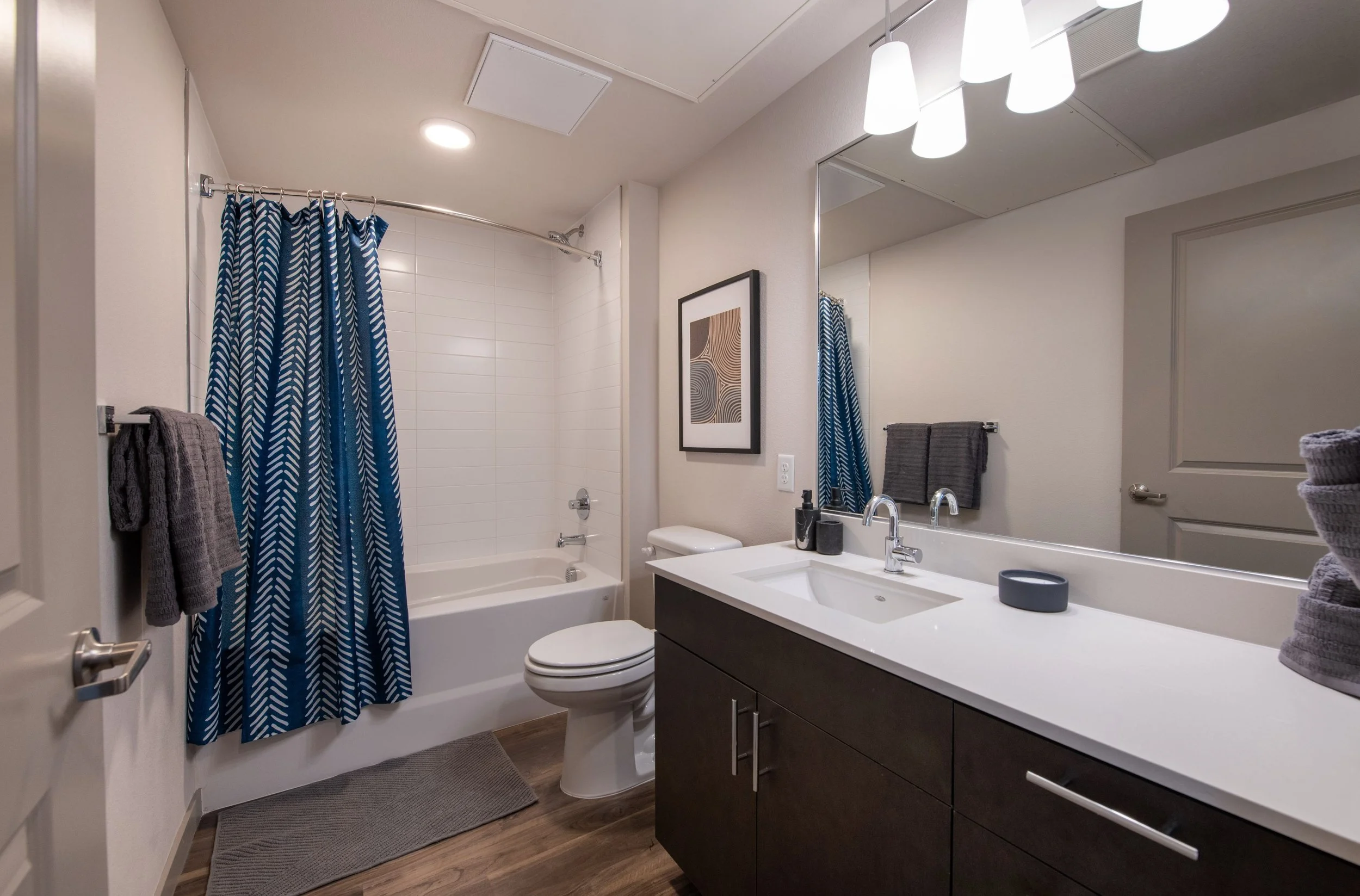 Modern bathroom with a white bathtub and shower, dark vanity with a white countertop, large mirror, framed artwork, towels, and geometric-patterned shower curtain in blue and white.