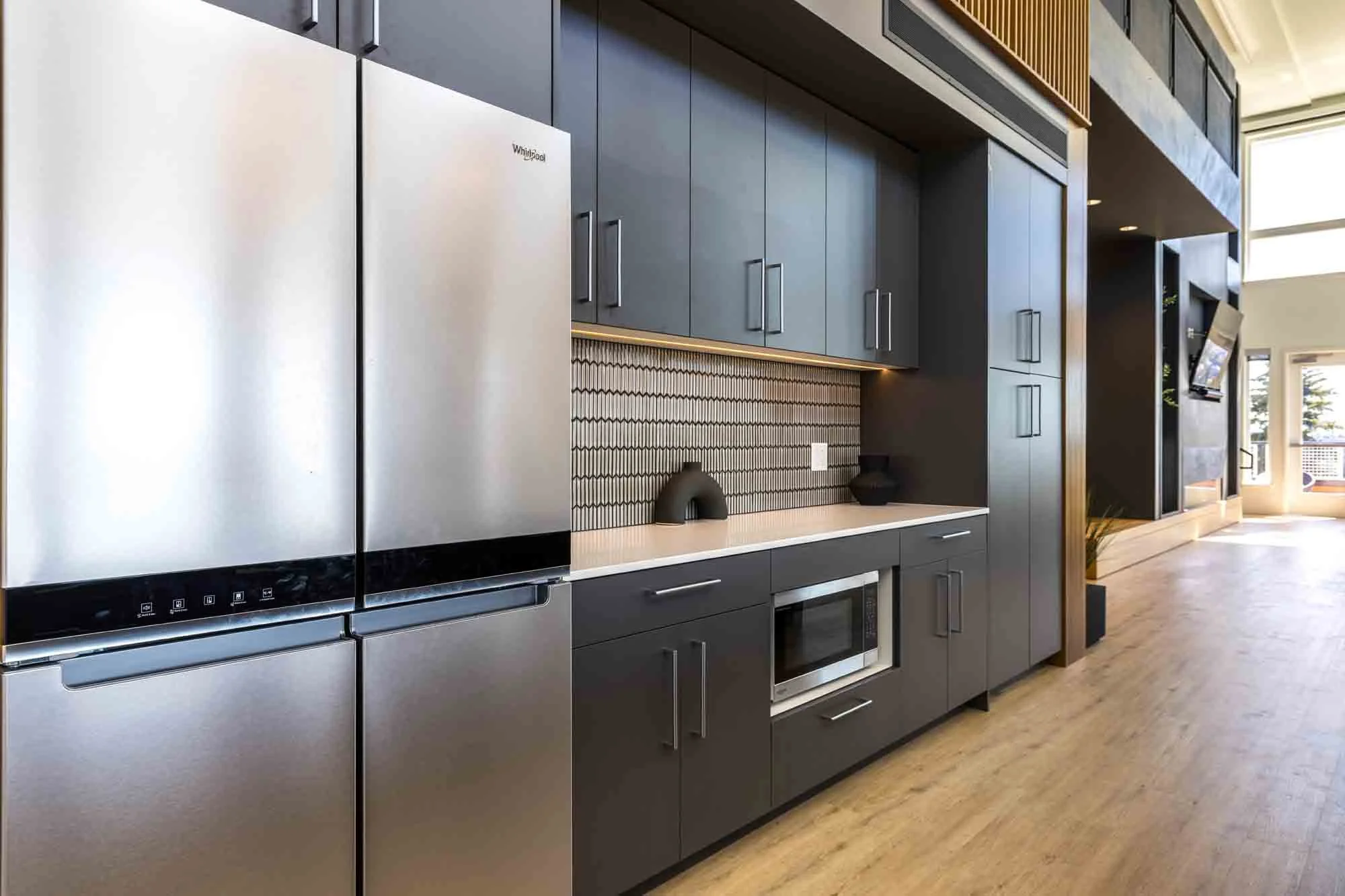Modern kitchen with gray cabinets, stainless steel refrigerator, microwave, and dark decorative backsplash, in a bright open space.