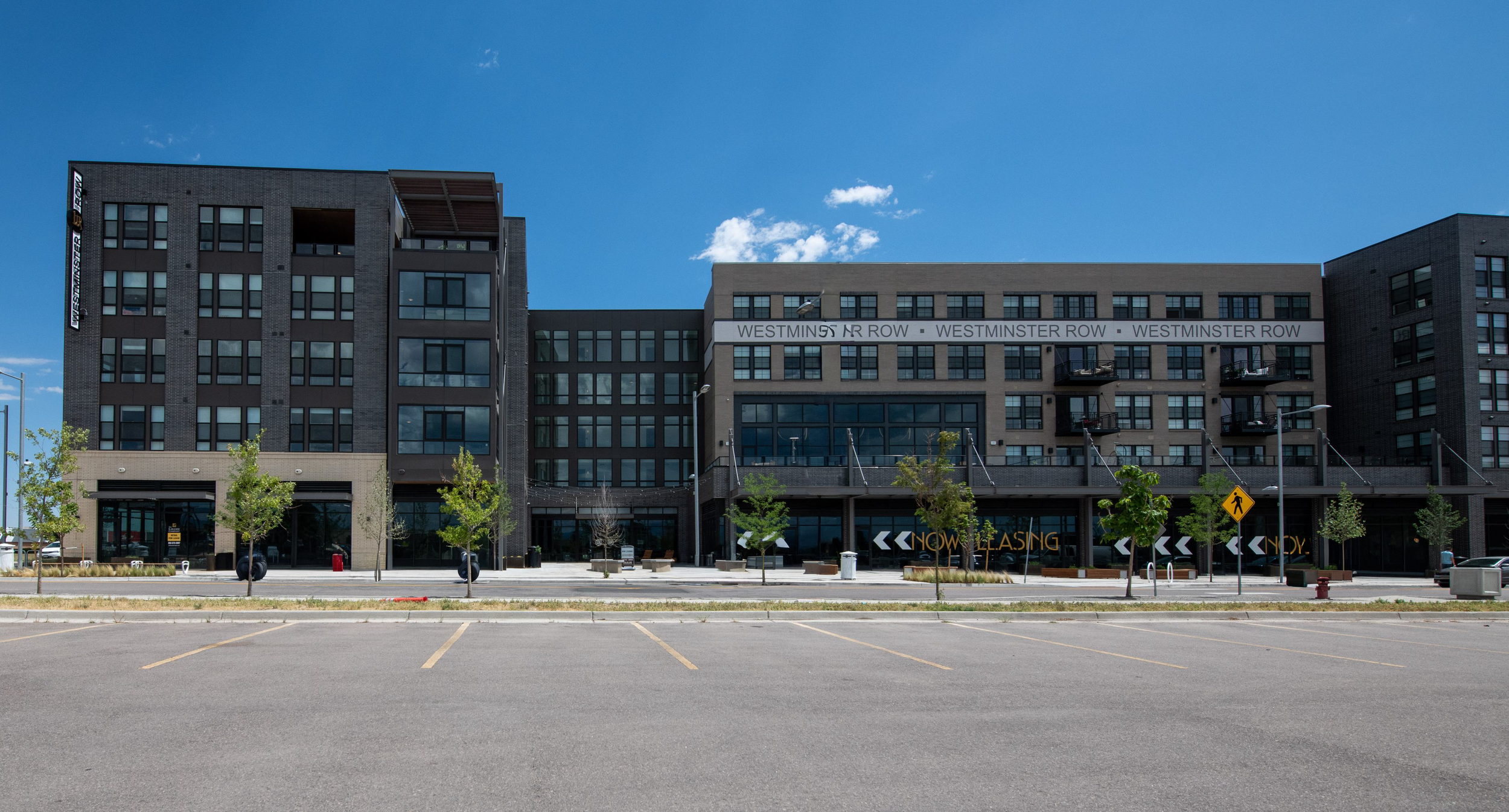 A modern multi-story building with a sign that reads "NOW LEASING" and "WESTMINSTER ROW" on the facade, with a mostly empty parking lot and a few small trees in the foreground under a clear blue sky.