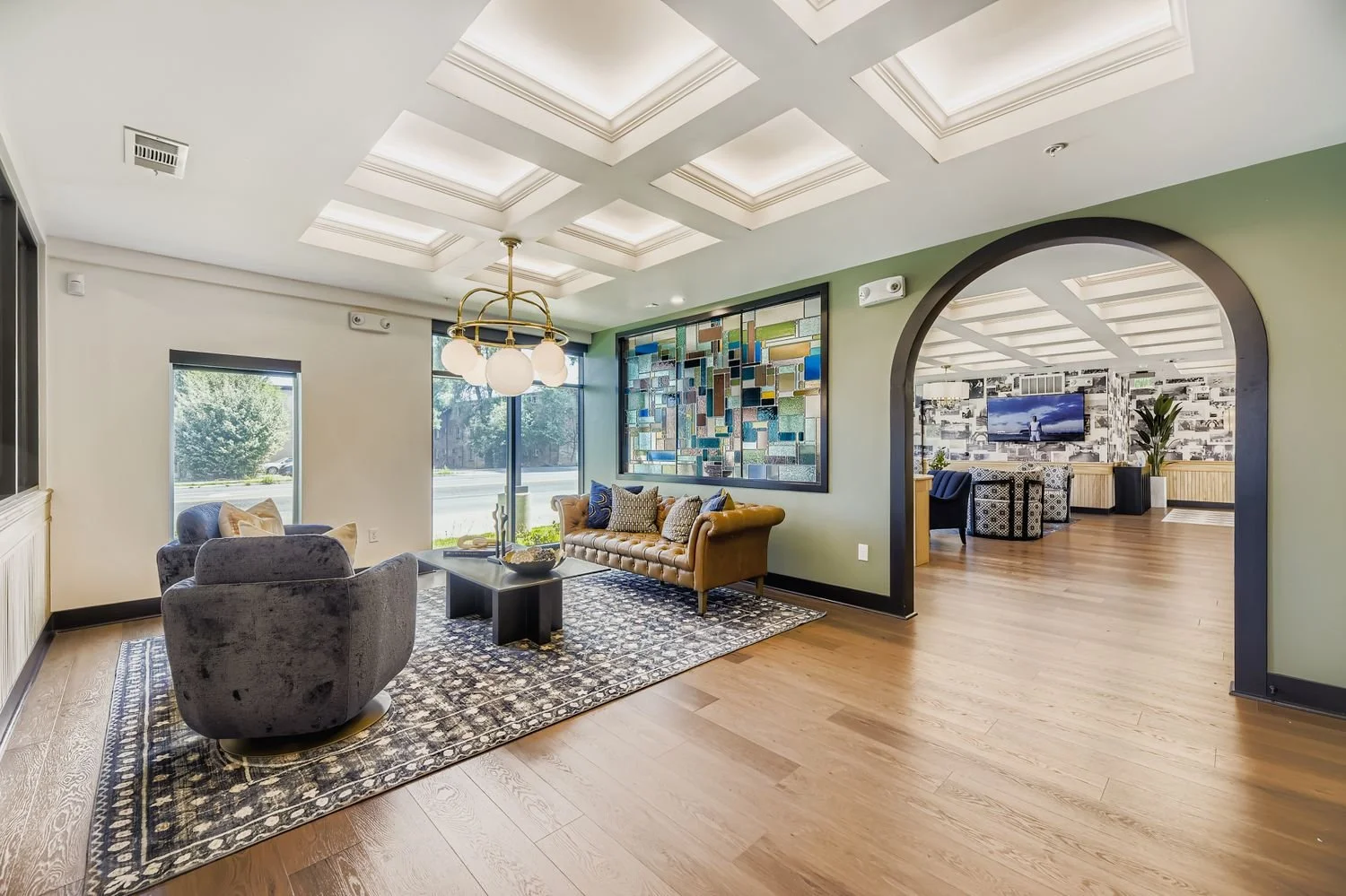 Living room with a tan leather sofa, gray armchairs, a black coffee table, and a decorative art piece on the wall. The room features wood flooring, a patterned rug, and large window with a view outside. An arched opening leads to a secondary room wit