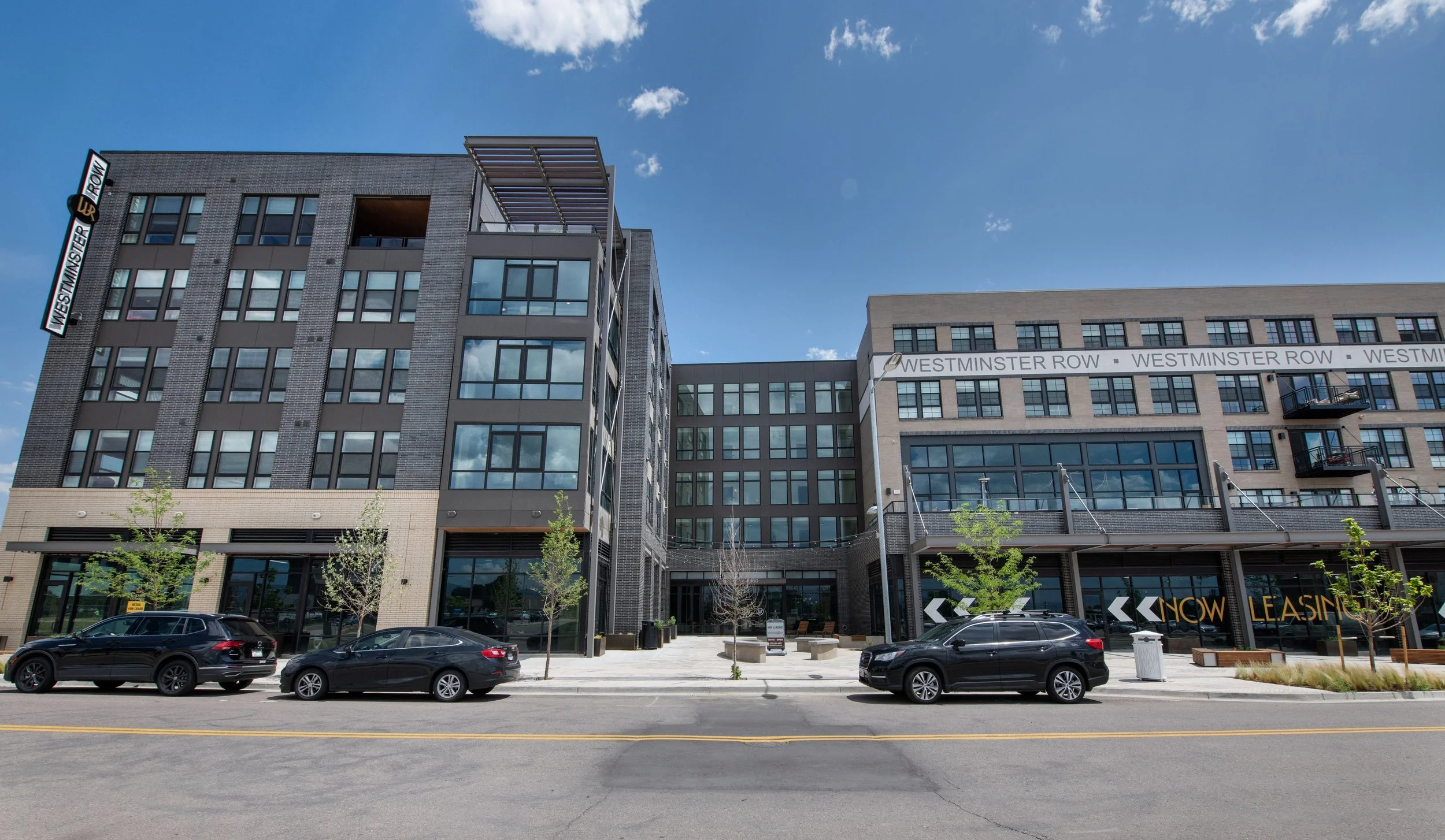 Modern multi-story apartment or office building with large windows, trees in front, parked cars on street, and a clear blue sky.
