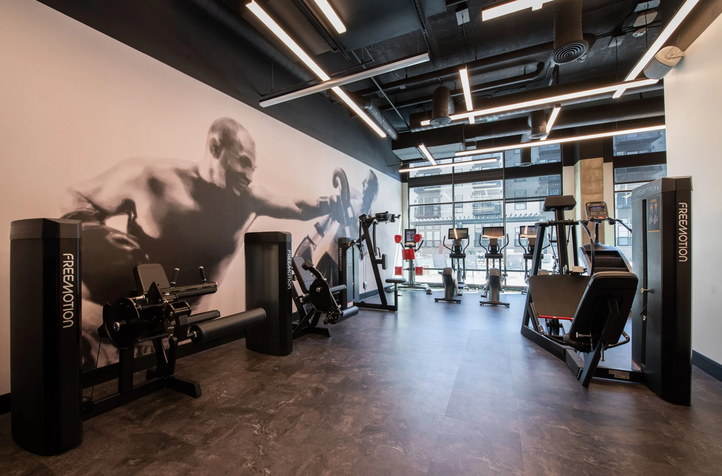 Modern gym with workout machines, a large black and white mural of a muscular man lifting weights on the wall, large windows showing city buildings, and stylish ceiling lights.