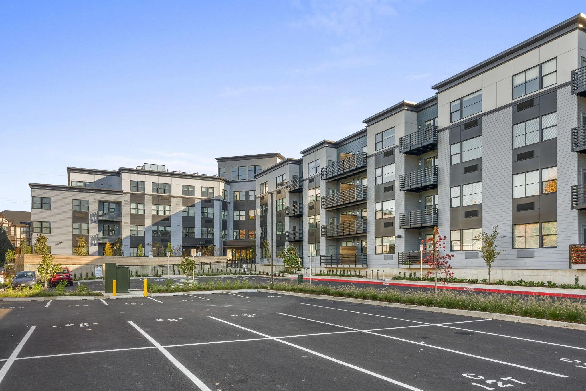 Modern multi-story residential apartment building with parking lot in the foreground, small trees, and landscaped areas, under a clear blue sky.