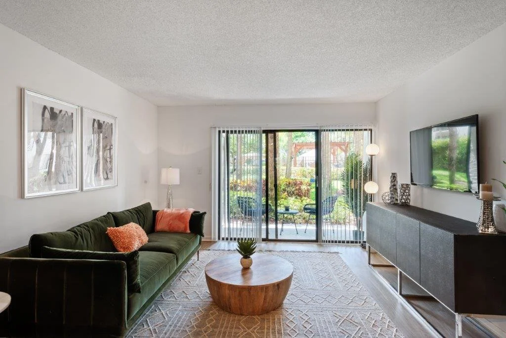 Living room with green sofa, round wooden coffee table, sliding glass doors with vertical blinds leading to a patio, wall-mounted flat-screen TV, and decorative items.