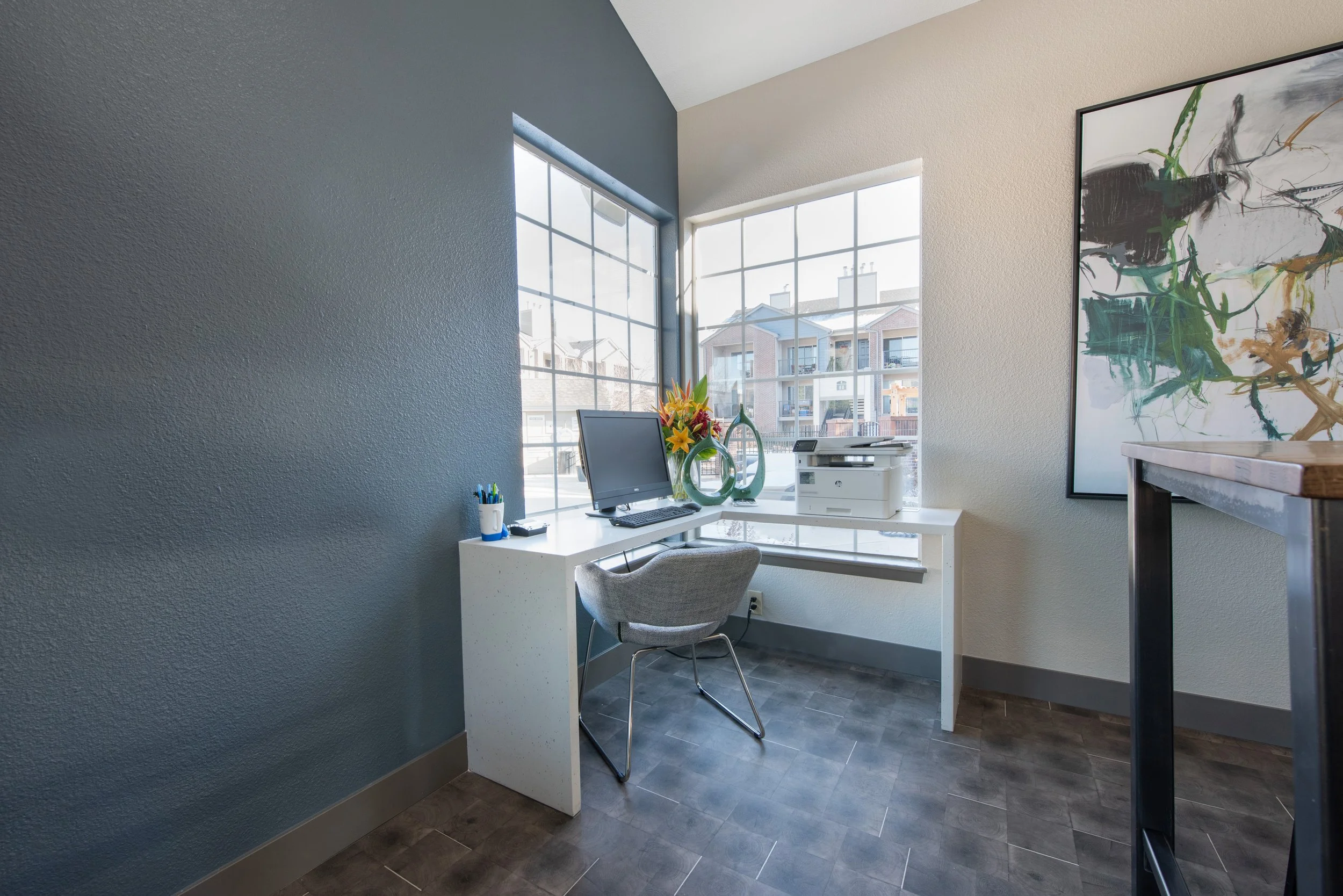 A small office space with a white desk in front of large windows, a computer monitor, fax machine, and decorative vases on the desk, a gray upholstered chair, a colorful abstract painting on the wall, and a dark wood high table to the right.