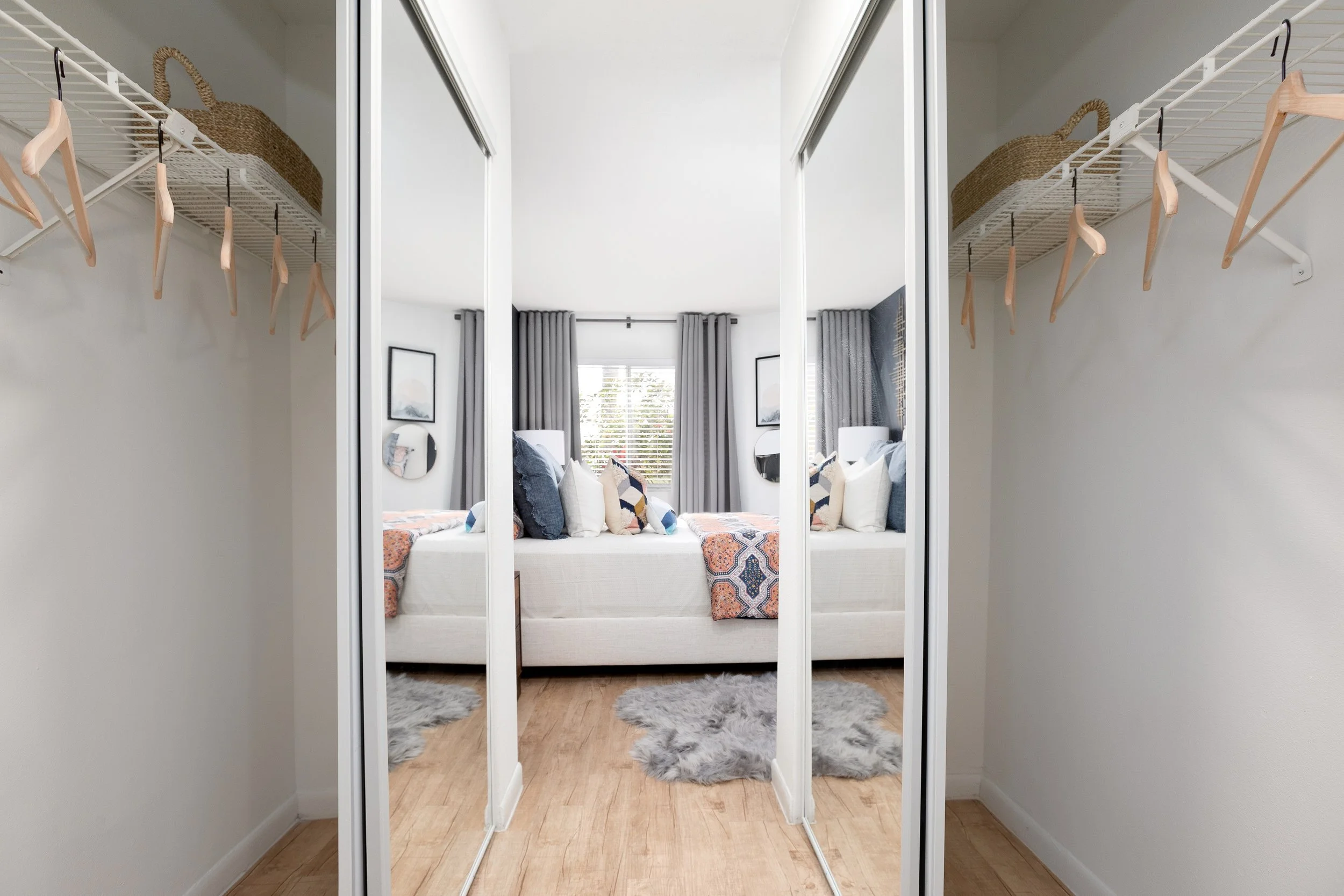 A mirror reflecting a cozy bedroom with a white bed, stacked pillows, and window with gray curtains.