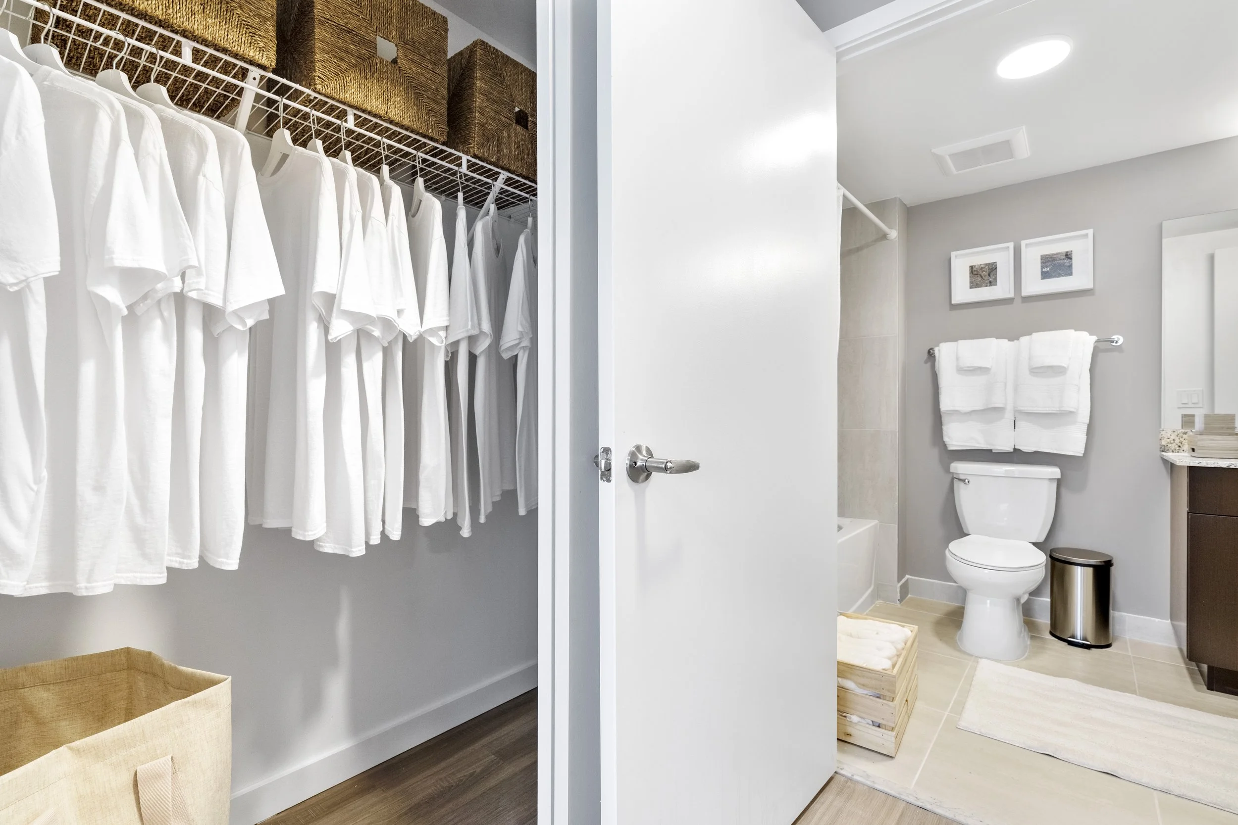 A walk-in closet with white shirts hanging on a shelf and baskets on top, opening into a bathroom with a toilet, towels, and framed pictures on the wall.