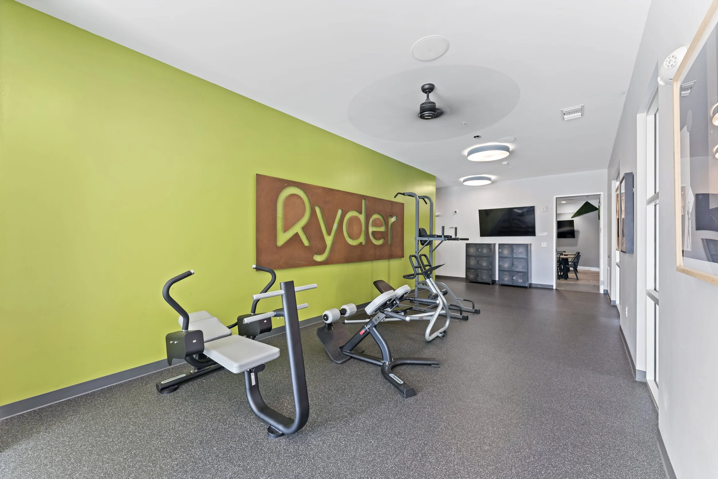 Indoor fitness room with exercise equipment, a green accent wall with the word "Ryder" on it, and a TV in the background.