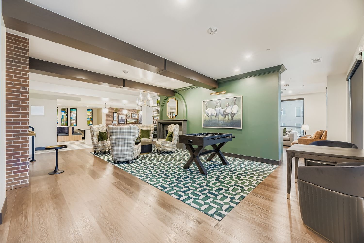 Interior of a lounge with a green accent wall, a framed painting of white herons, a foosball table, and comfortable seating including checkered and beige armchairs, with a patterned rug and wooden flooring.