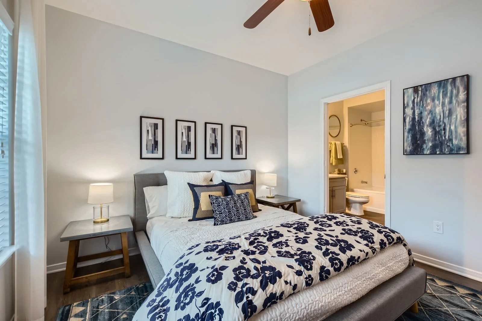 Modern bedroom with a bed featuring a navy and white floral comforter, multiple pillows, and nightstands with lamps. There are four abstract art pieces above the bed and an artwork on the wall. An en suite bathroom is visible through an open door.