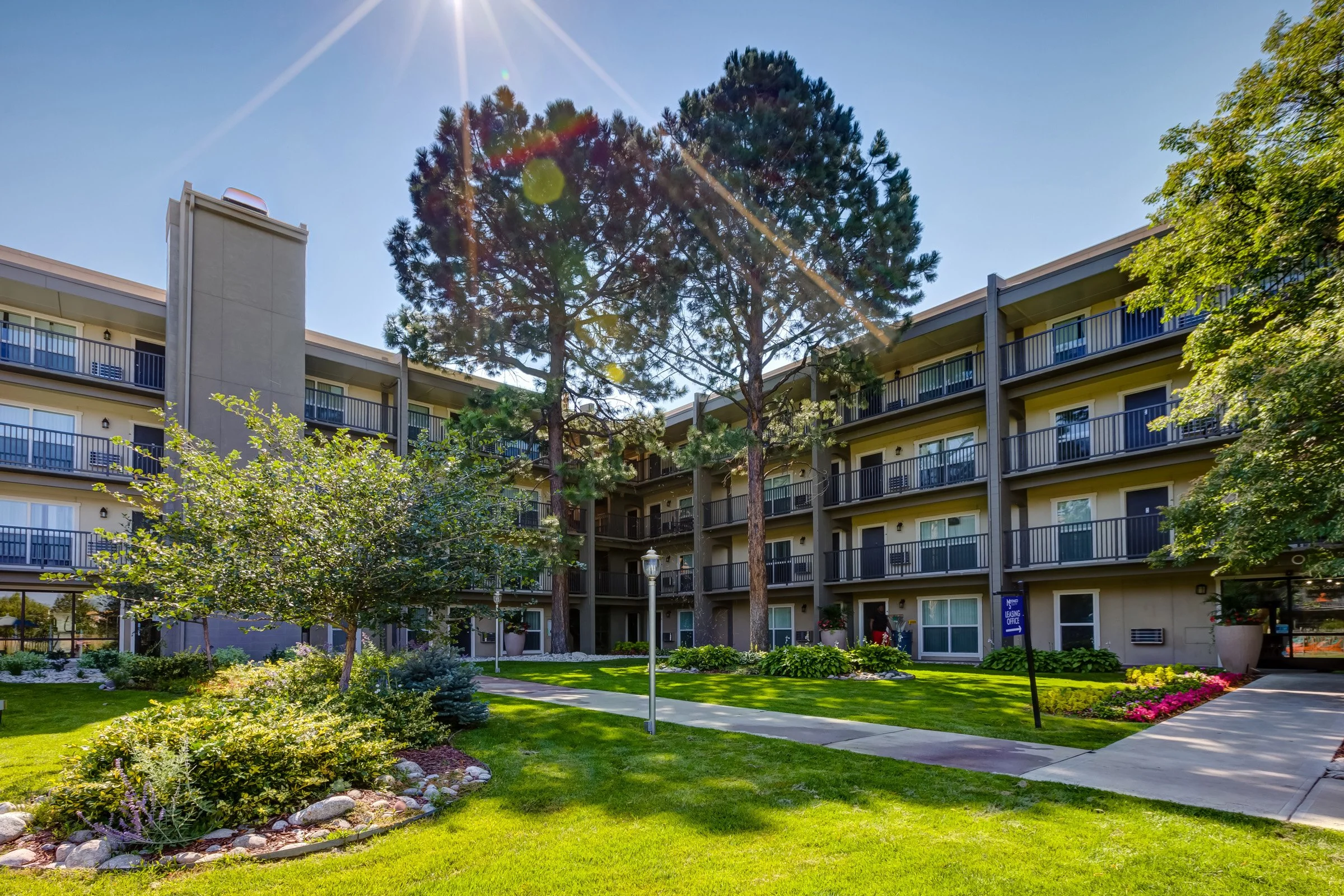 A multi-story apartment building with balconies, surrounded by a landscaped garden with trees, shrubs, and a walking path, under a sunny sky with lens flare.