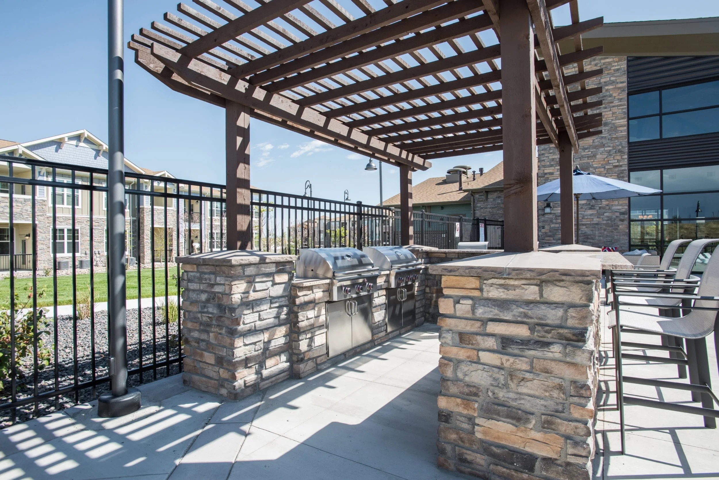 Outdoor barbecue area with two stainless steel grills built into stone and brick countertops, wooden pergola overhead, surrounded by black metal fence, with patio chairs and an umbrella in the background.