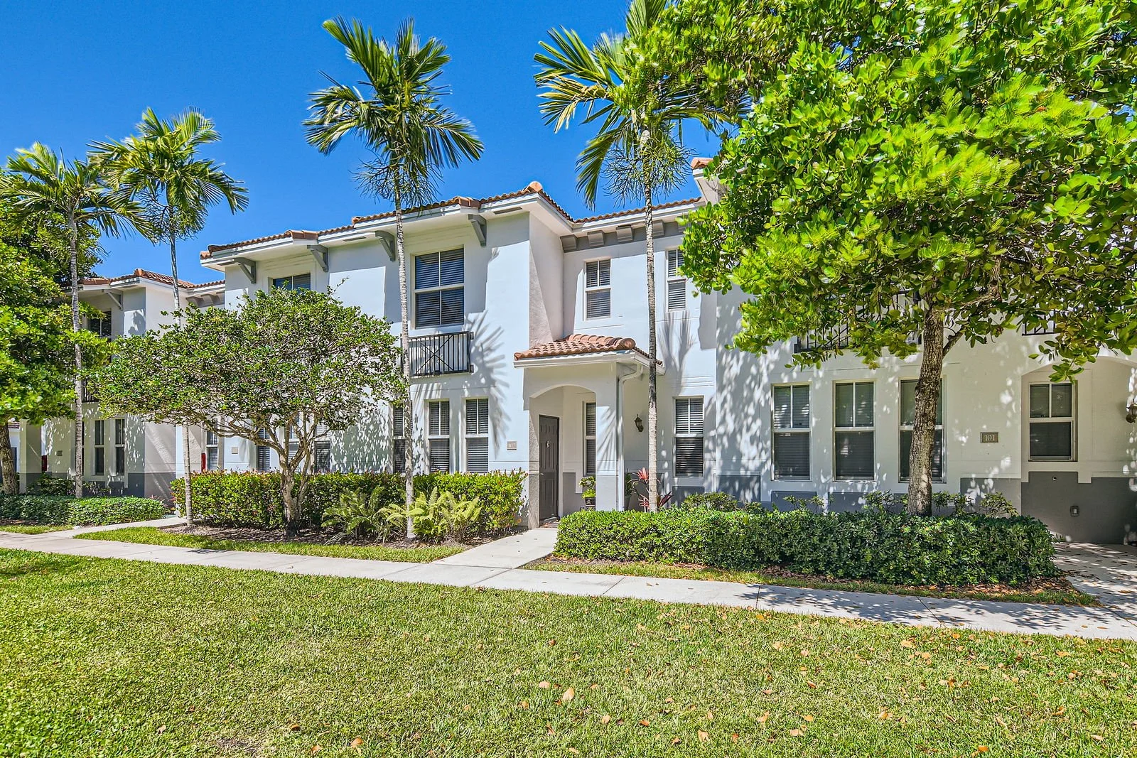 White multi-story residential building with terracotta tile roof, surrounded by green trees and bushes, on a sunny day.