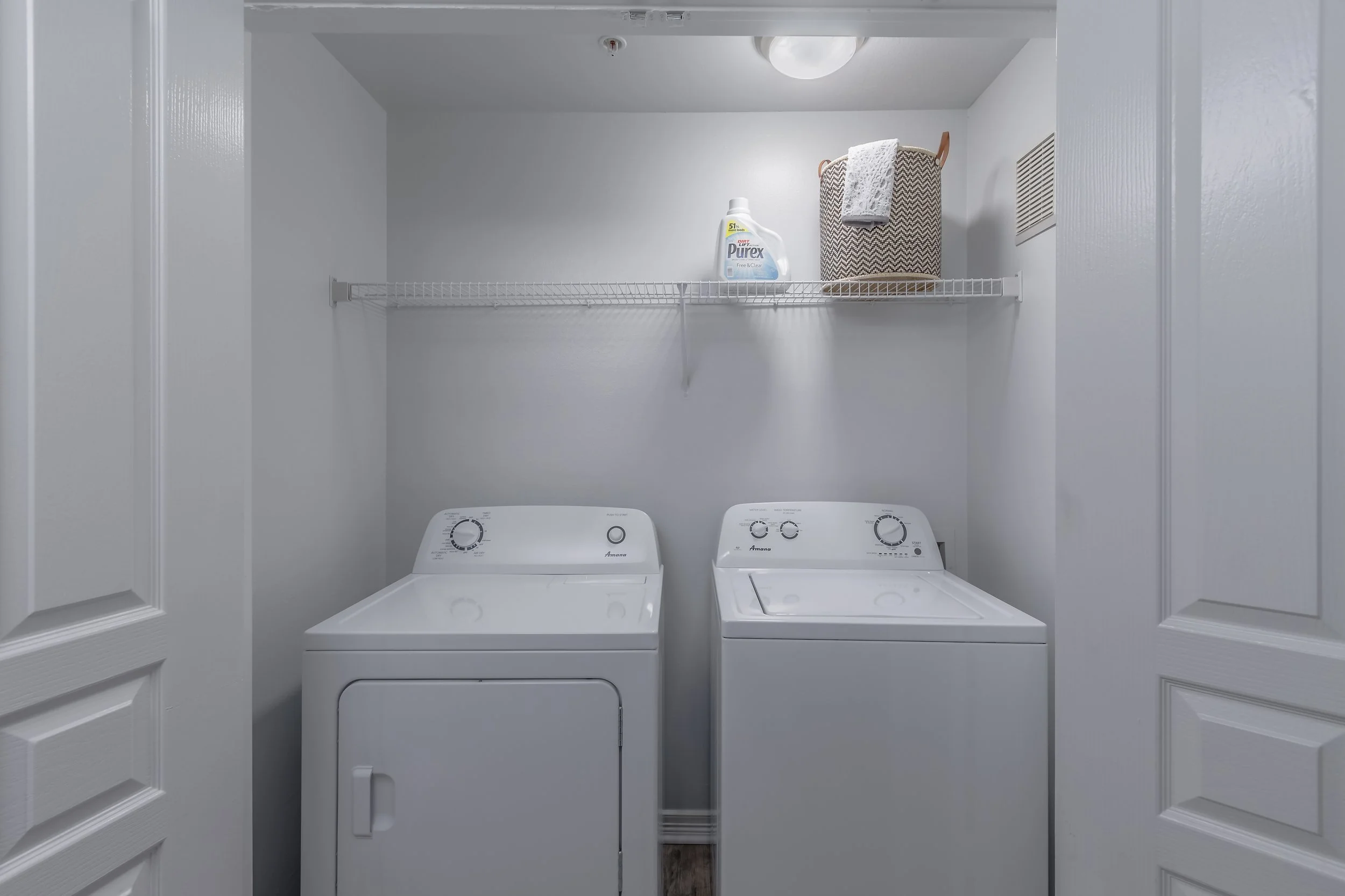 A laundry room with a white washer and dryer side by side, a wire shelf above holding a detergent bottle, a basket with a towel, and a vent on the right wall.