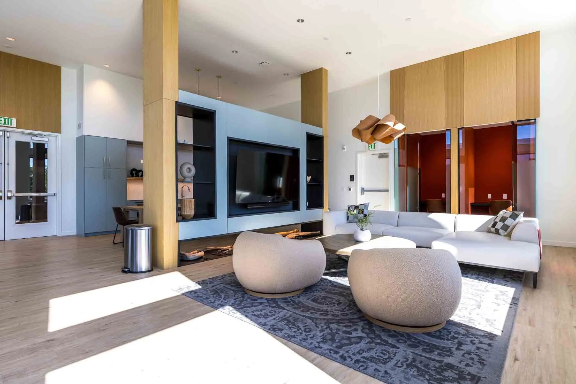 Modern living room with a large white sectional sofa, two rounded beige chairs on a patterned rug, a black coffee table with a plant, a large screen TV on a blue wall unit, and a wooden ceiling pendant light.