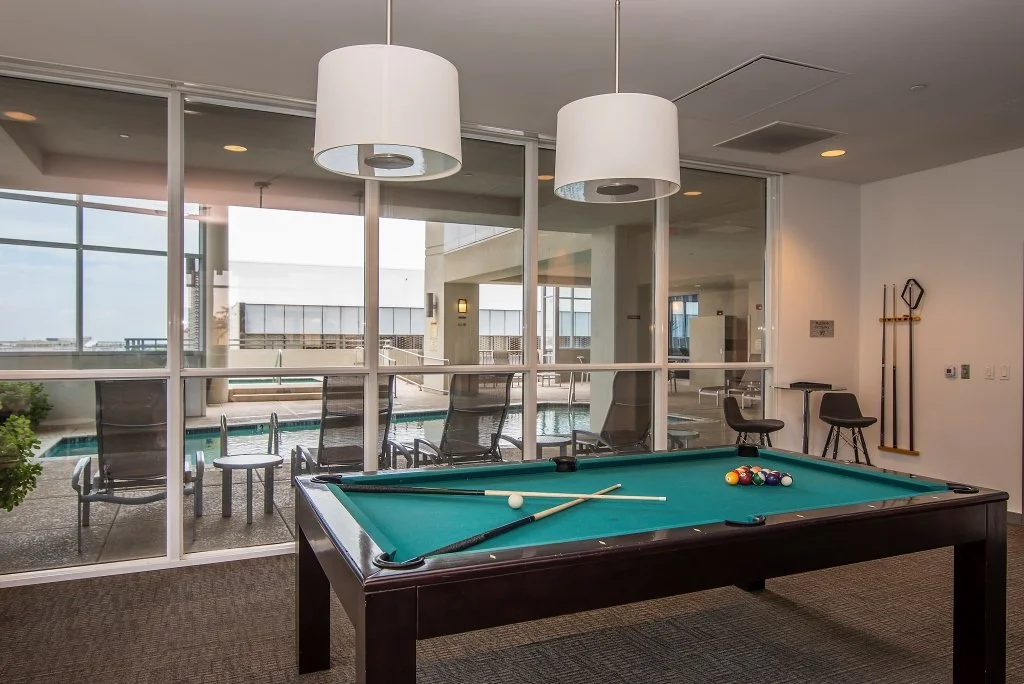 Indoor recreational room with a pool table, pool balls, cue sticks, and a view of an outdoor pool area through large glass windows.