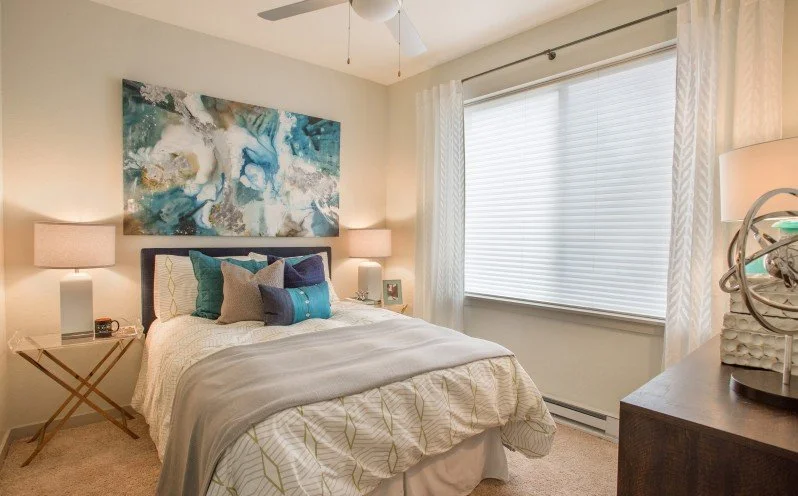 A bedroom with a bed, multiple pillows, two bedside lamps, a large abstract painting above the headboard, a window with blinds, and a dresser with a decorative object on top.