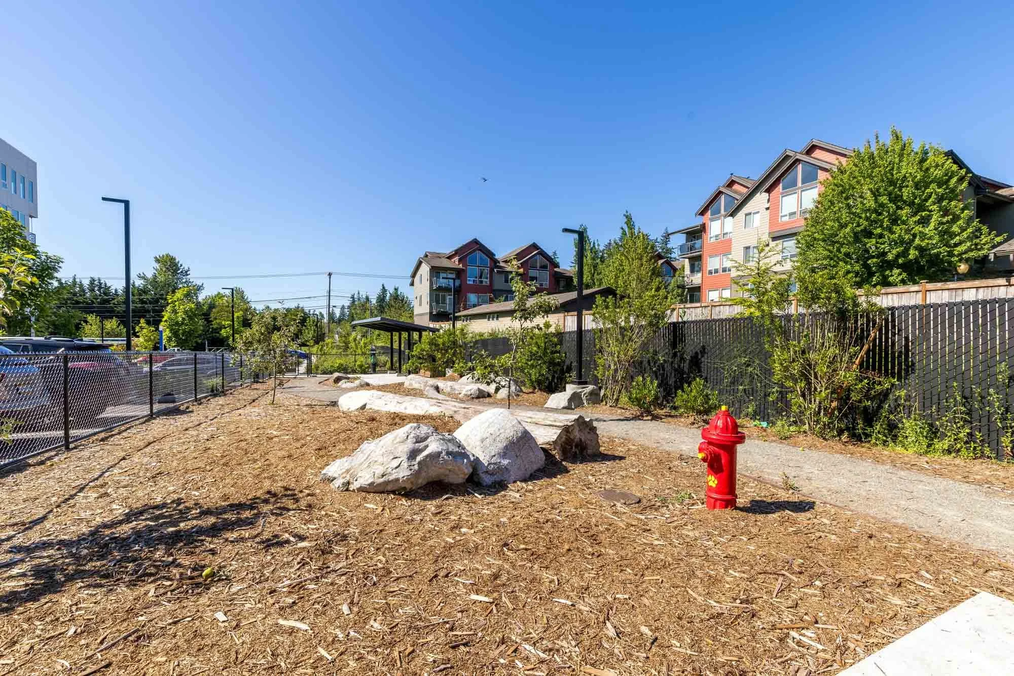 A landscaped urban park with mulch ground, large rocks, trees, a red fire hydrant, and a paved walkway, surrounded by residential buildings and a black fence under a clear blue sky.