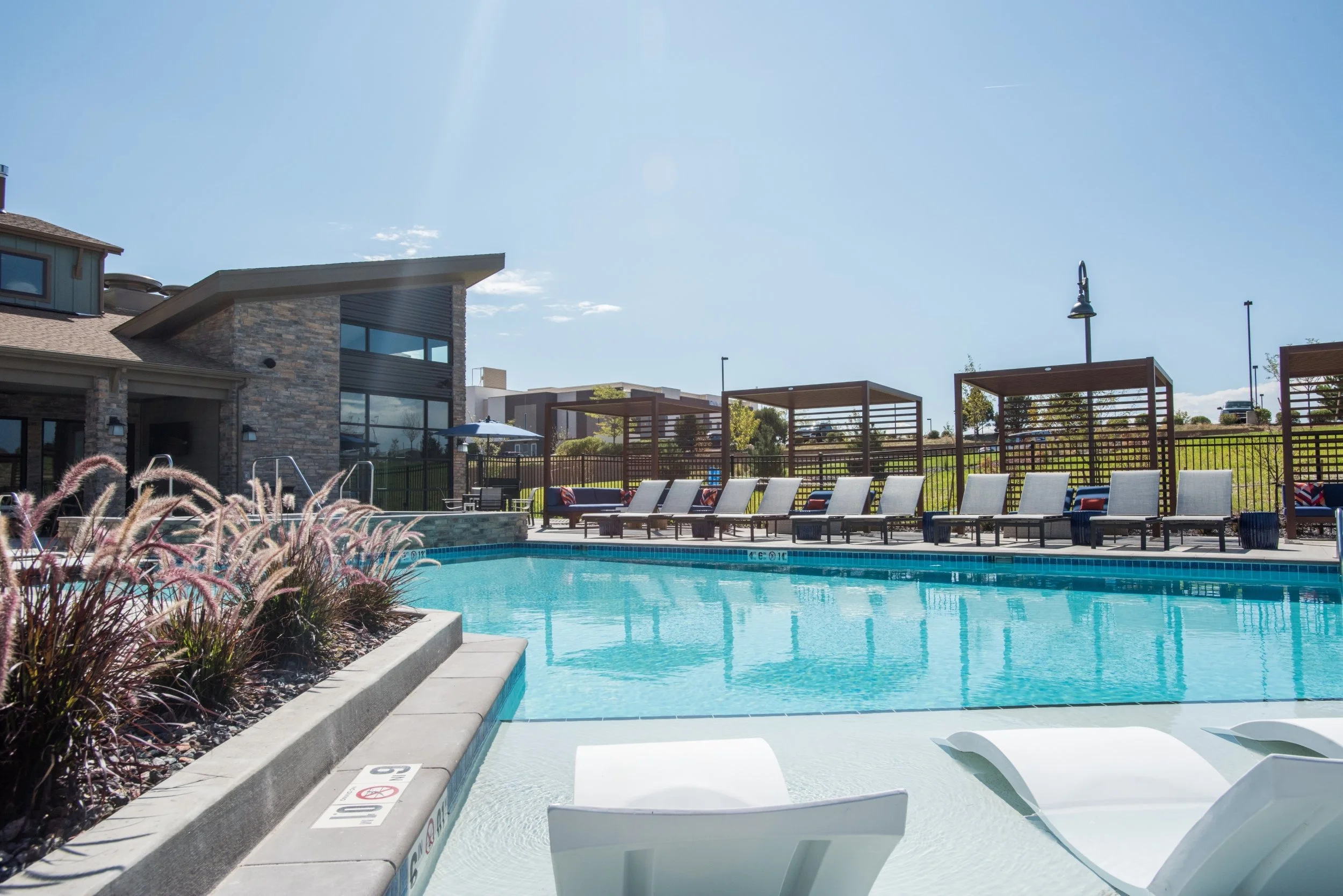 Empty outdoor swimming pool with lounge chairs, cabanas, and a modern residential building under a clear blue sky.