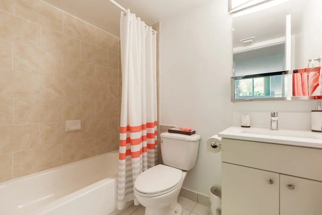 Bathroom with bathtub, beige tiles, white toilet, small vanity with sink, mirror, and red accents.