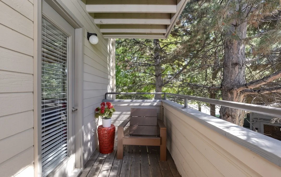 Small balcony with a wooden chair, a red pot with pink flowers, beige siding, a glass door with blinds, and a tree with green leaves in the background.