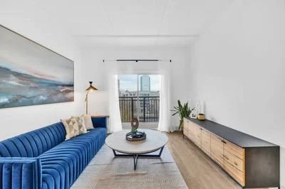 Living room with a blue velvet sofa, a round coffee table, a large abstract painting, a wooden TV stand, a tall lamp, and a balcony with city view.