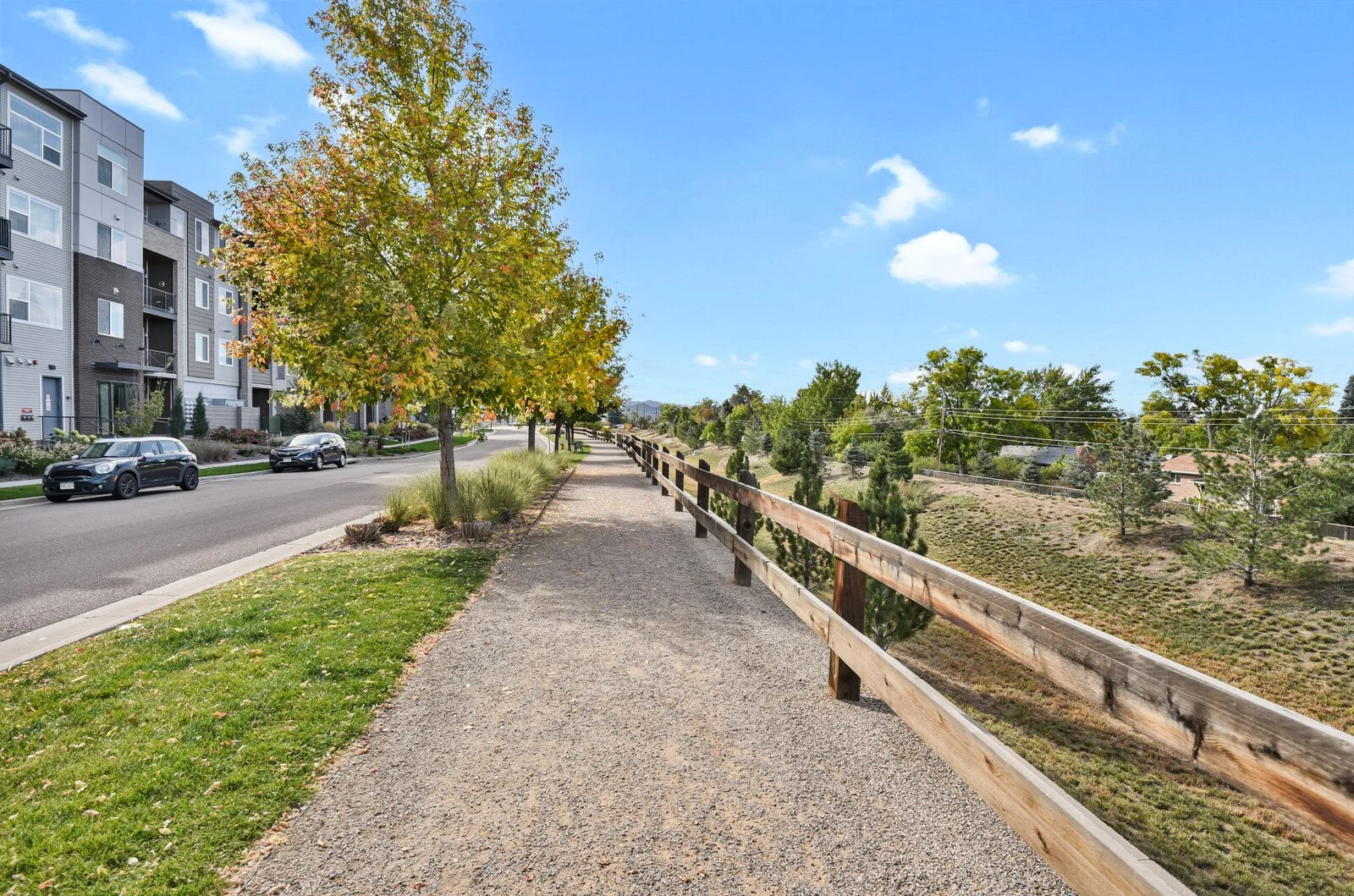 A sidewalk runs parallel to a street with parked cars on the left; a tree with green and yellow leaves is beside the sidewalk, and a wooden railing separates the sidewalk from a grassy slope with small trees and shrubs; overhead, power lines are visi
