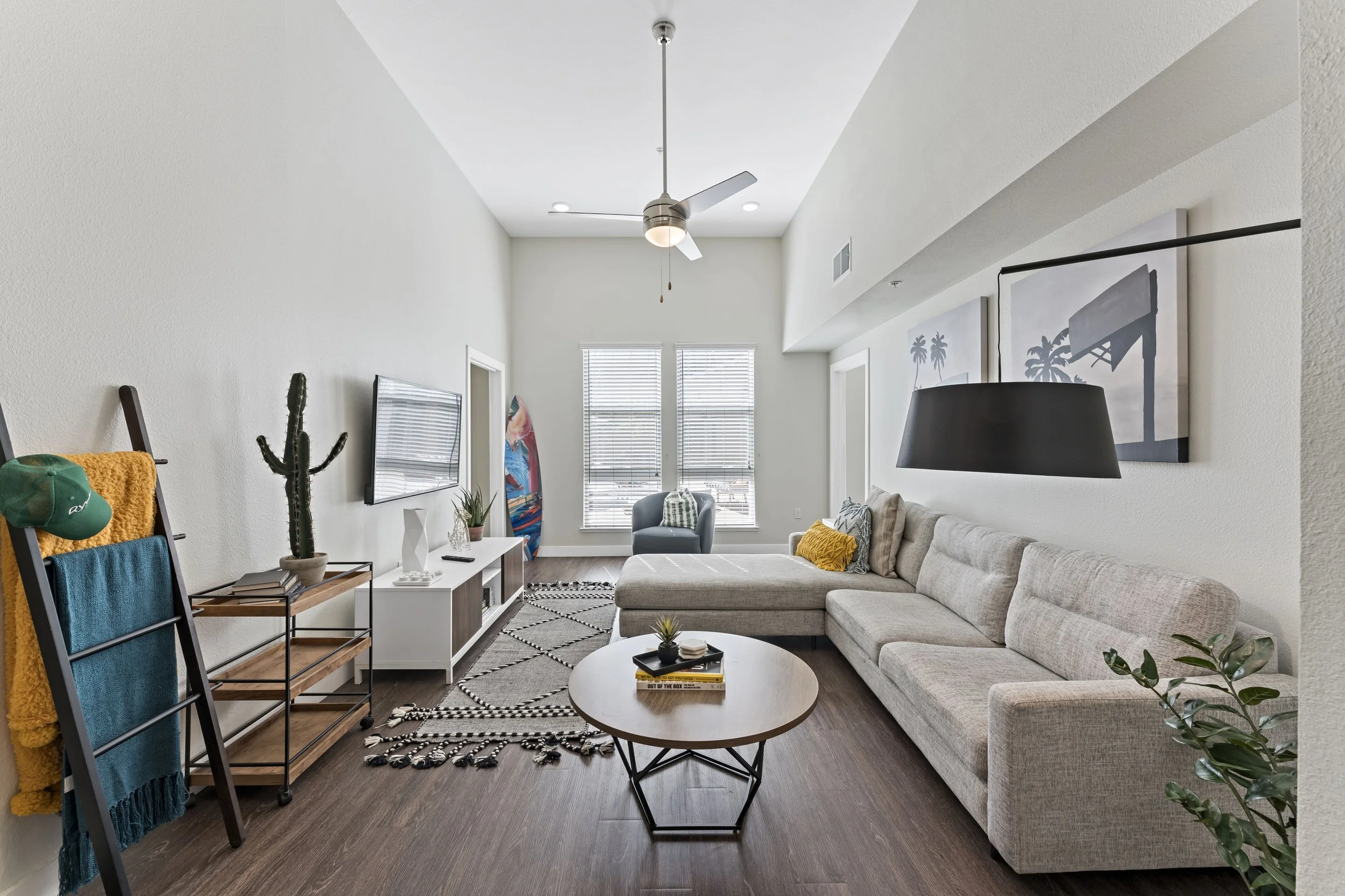 Living room with beige sectional sofa, round coffee table, black pendant light, television on white stand, decorative plants, and wall art of palm trees and a surfboard, with hardwood floors and white walls.
