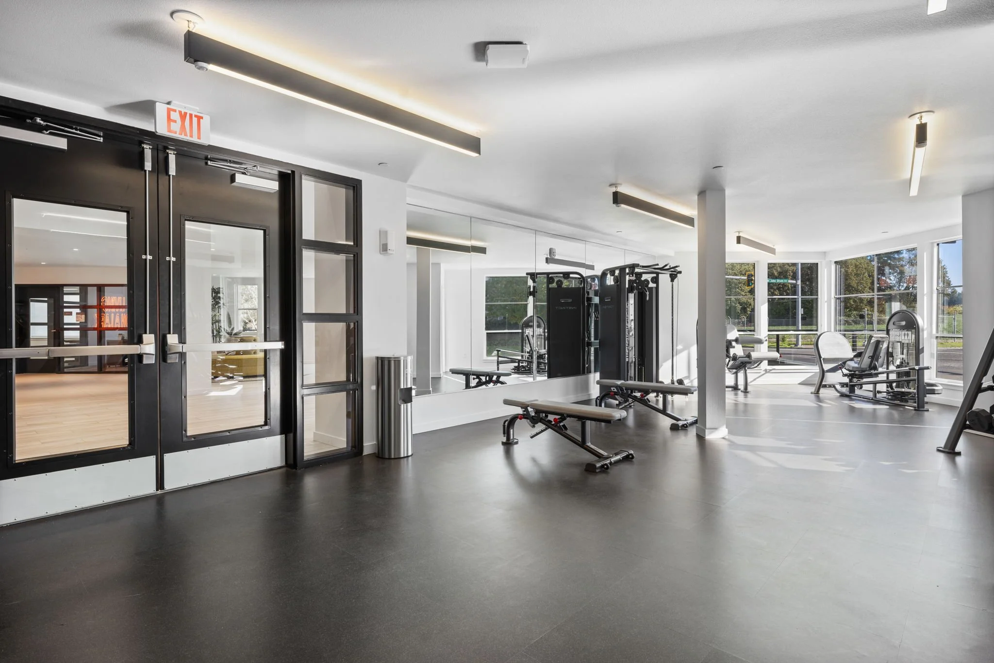 Modern gym interior with workout equipment including weight machines, benches, and exercise chairs, large windows allowing natural light, and black double doors with an exit sign above.