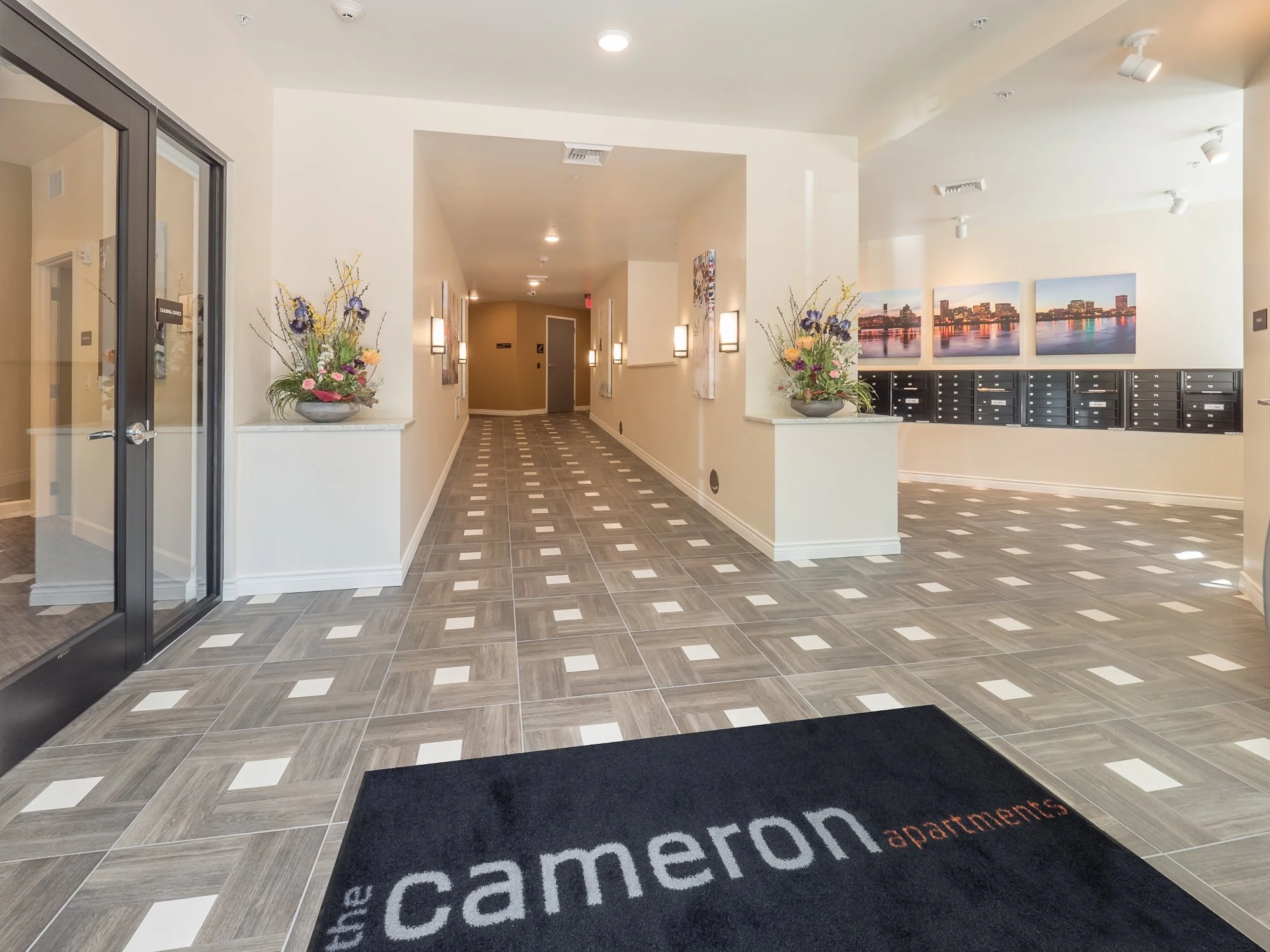 Lobby of The Cameron Apartments with tiled floor, flower arrangements on ledges, mailboxes on wall, and framed cityscape photos.