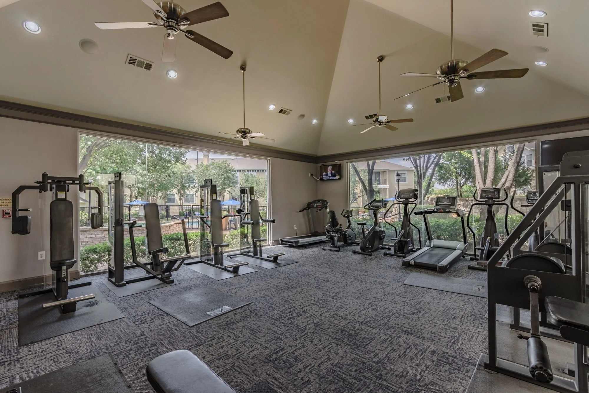 Indoor gym with exercise equipment including weight machines, treadmills, and stationary bikes, with large windows showing outdoor trees and buildings.