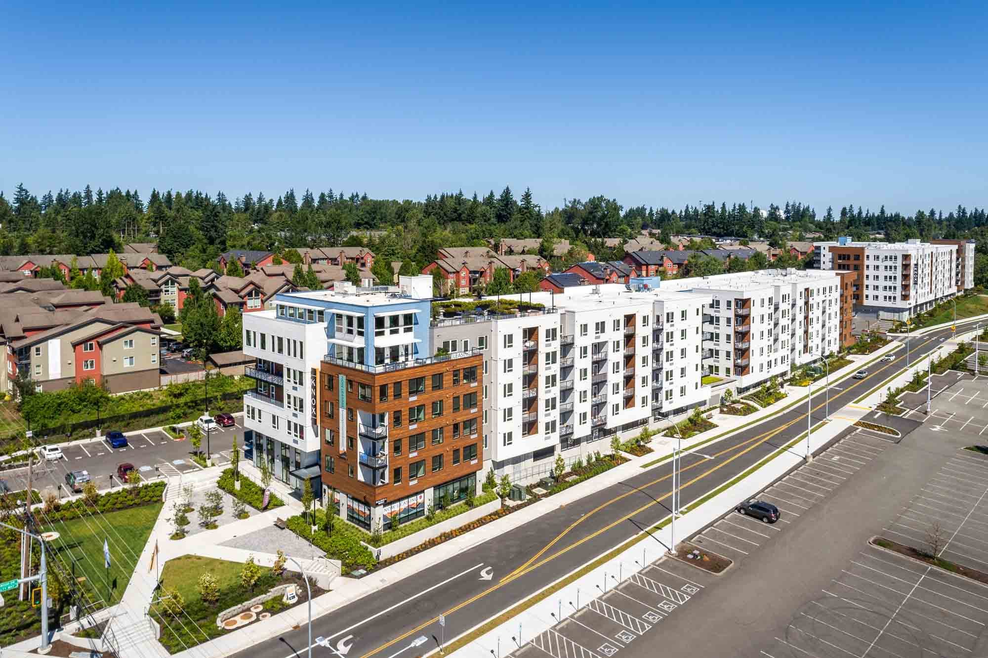 Aerial view of a modern apartment complex with multiple white and brown buildings, surrounded by parking lots, trees, and a residential neighborhood with single-family homes in the background, under a clear blue sky.