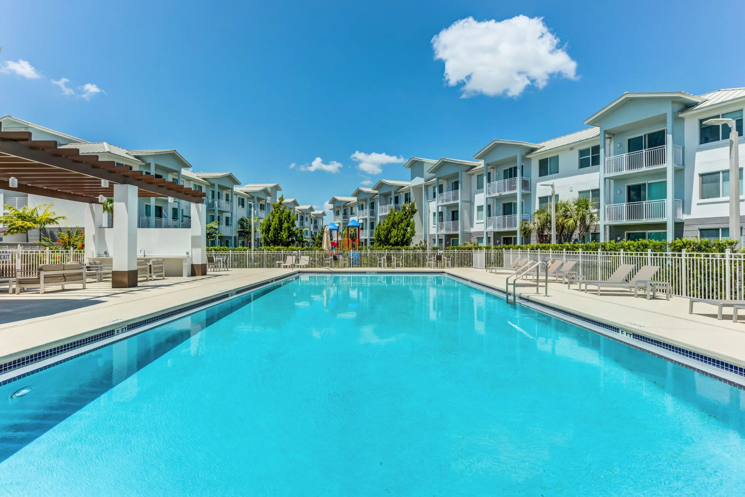 An outdoor swimming pool area in an apartment complex with blue sky and white puffy clouds, surrounded by multi-story residential buildings with balconies.