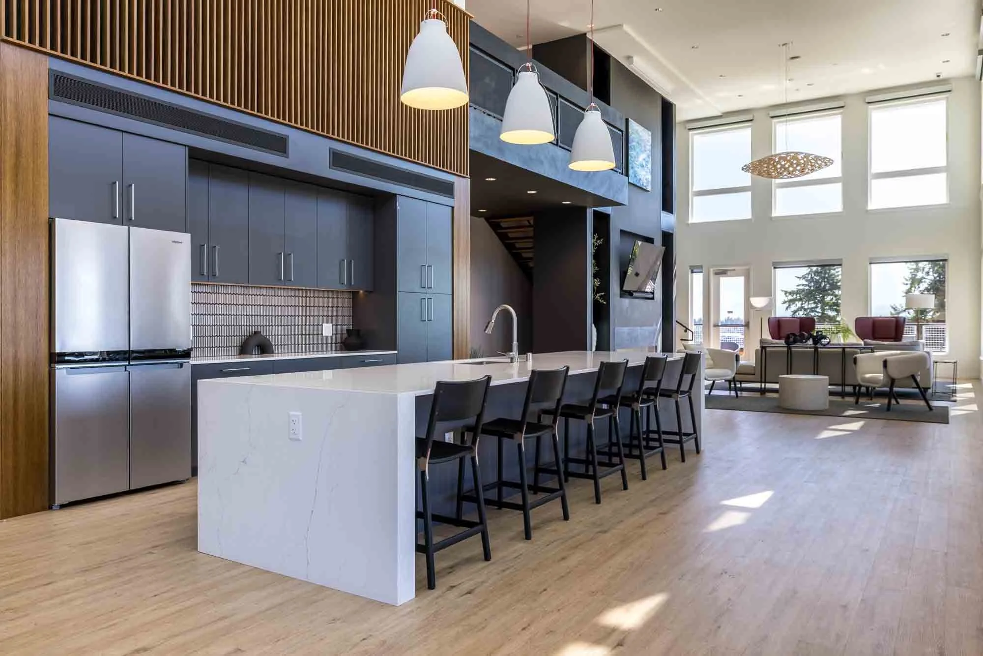 Modern open-concept kitchen and lounge area with large windows, featuring a white marble kitchen island, dark cabinetry, a stainless steel refrigerator, and seating for six at the island. The lounge space has white and red chairs, a coffee table, and