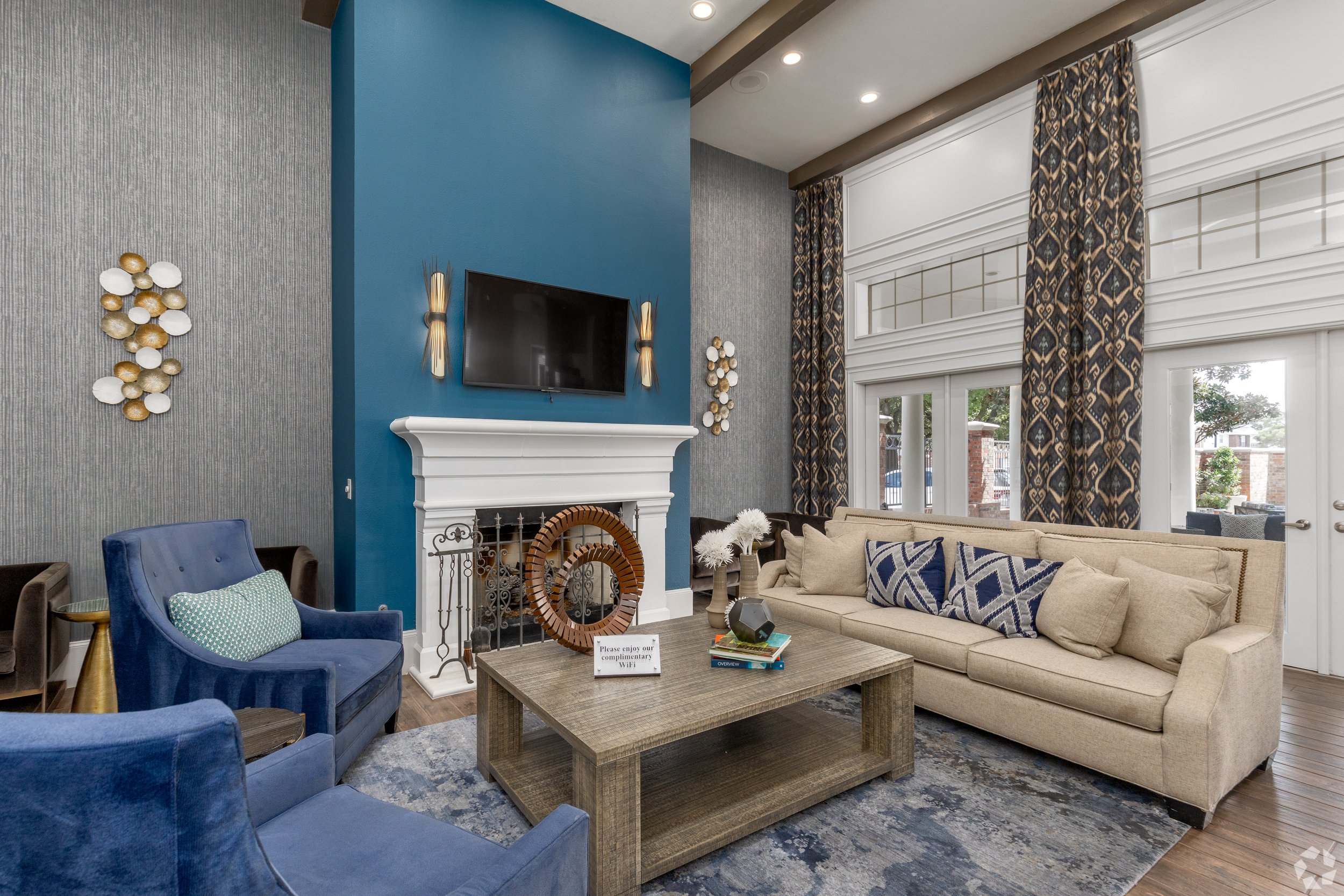 Living room with a beige couch, blue armchairs, a wooden coffee table, a white fireplace, a wall-mounted TV, large windows with patterned curtains, and modern wall art.