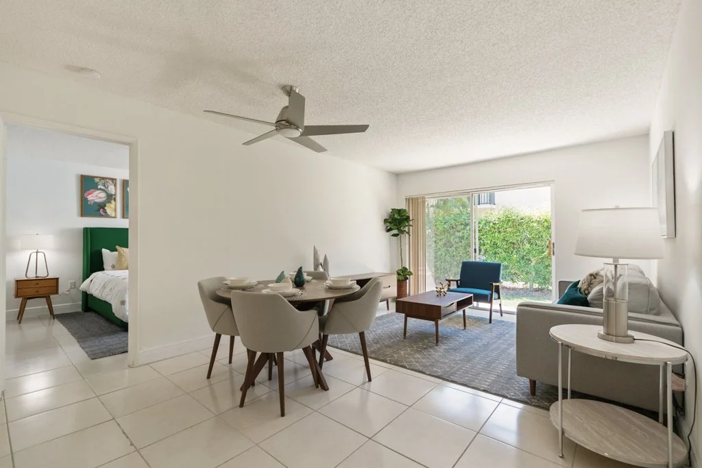 Modern living room with dining area, white walls, tile flooring, sliding glass door with greenery outside, sofa, armchair, coffee table, side table with lamp, ceiling fan, and a view into a bedroom with green headboard.