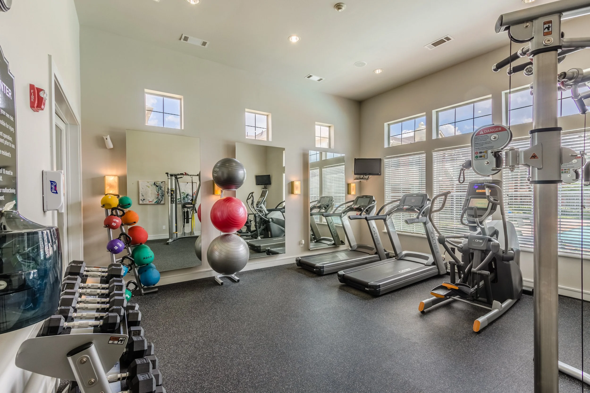 A clean, well-lit gym with various exercise equipment including treadmills, a stationary bike, and elliptical trainers. The room has large windows with blinds, a mirror, and a small set of colorful medicine balls and dumbbells.