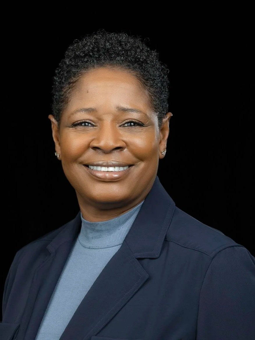 Portrait of a smiling African American woman with short curly hair, wearing a dark blazer and light blue turtleneck, against a black background.
