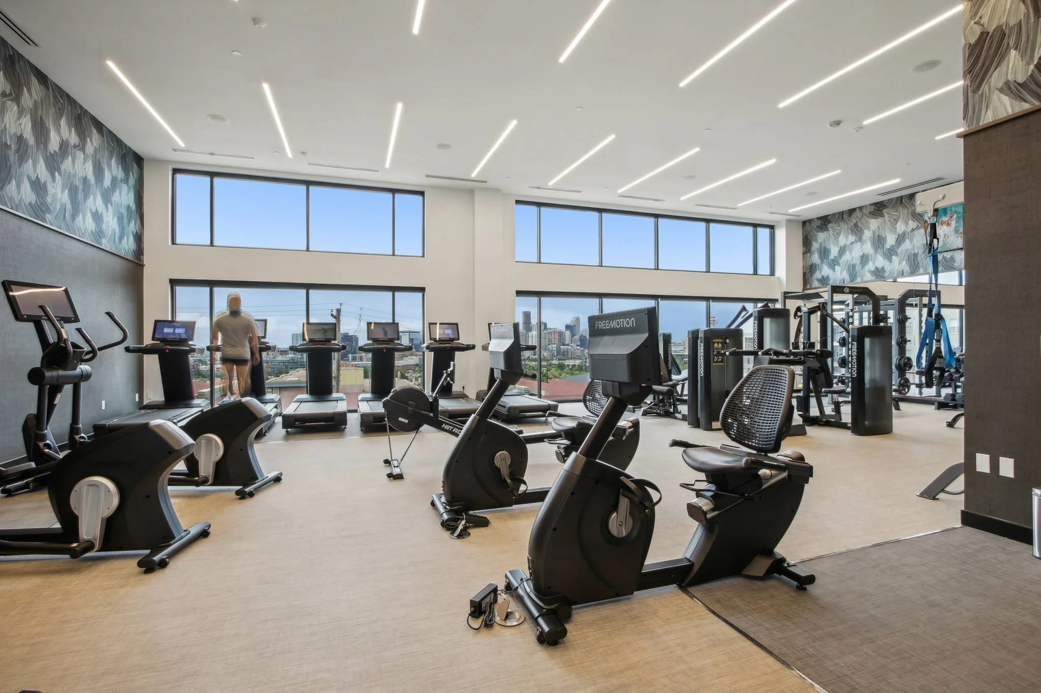 Modern gym with cardio equipment including treadmills, stationary bikes, and elliptical machines, large windows with city view, and a person working out