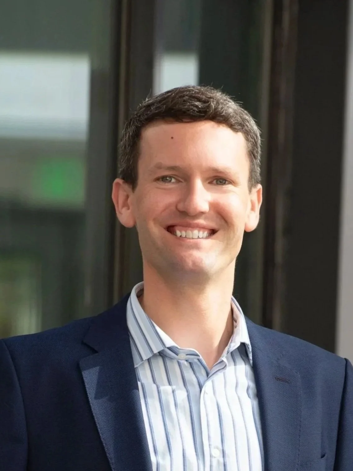 A smiling man with short brown hair wearing a blue blazer and a light blue striped shirt, posing indoors in front of a window.