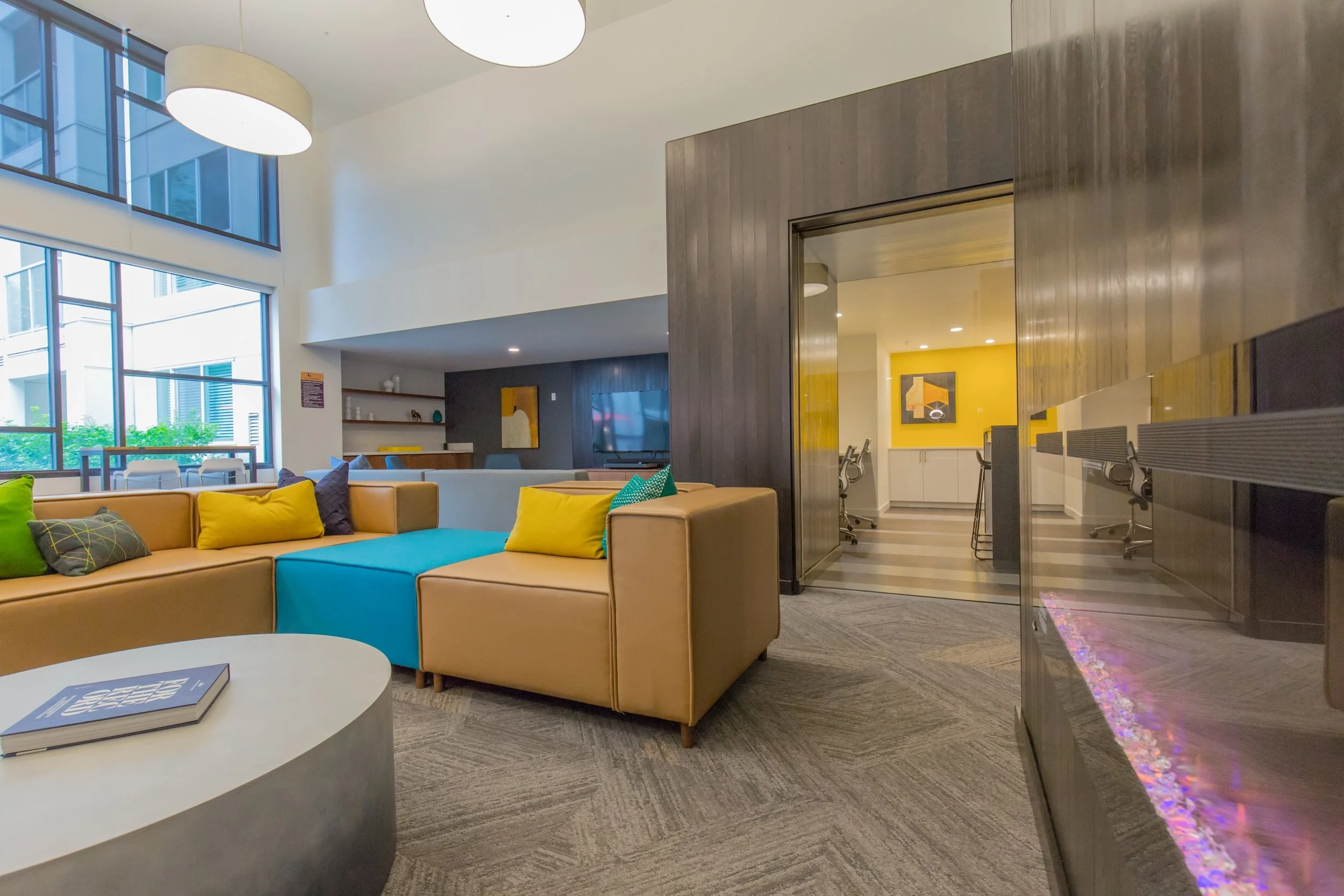 Modern lobby with large windows, tan sectional sofa with colorful pillows, a round coffee table with a book, a fireplace with purple lighting, and a meeting room with yellow accent wall and chairs.
