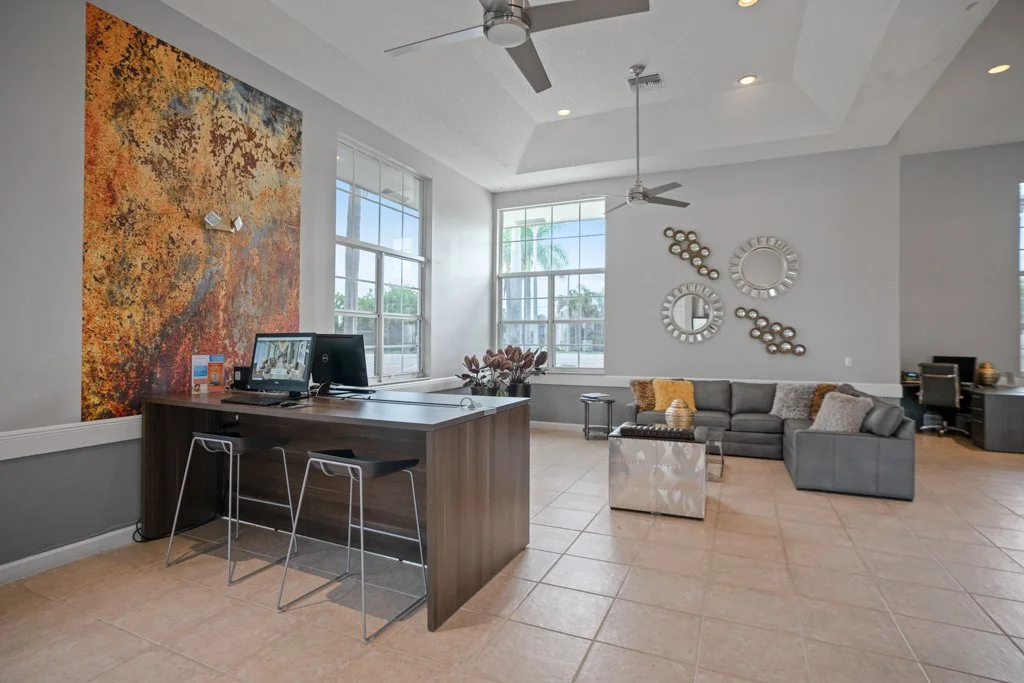 Modern living room with large windows, a gray sectional sofa, circular decorative mirrors on the wall, a desk with two chairs, and a colorful abstract wall art.