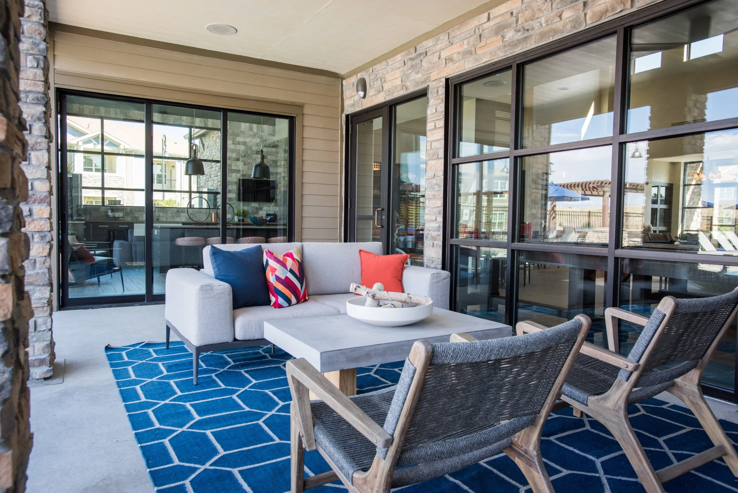 Outdoor patio area with a light gray sofa, colorful pillows, a wooden armchair, a blue patterned rug, and a concrete coffee table with decorative items. The background features large glass sliding doors and windows showing the interior of a modern ho