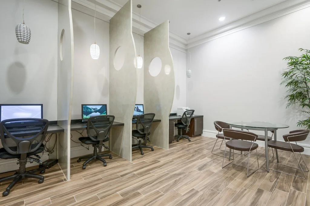 Office space with three computer stations separated by decorative dividers, a round glass table with four chairs, and a tall potted plant, featuring wooden flooring and hanging pendant lights.
