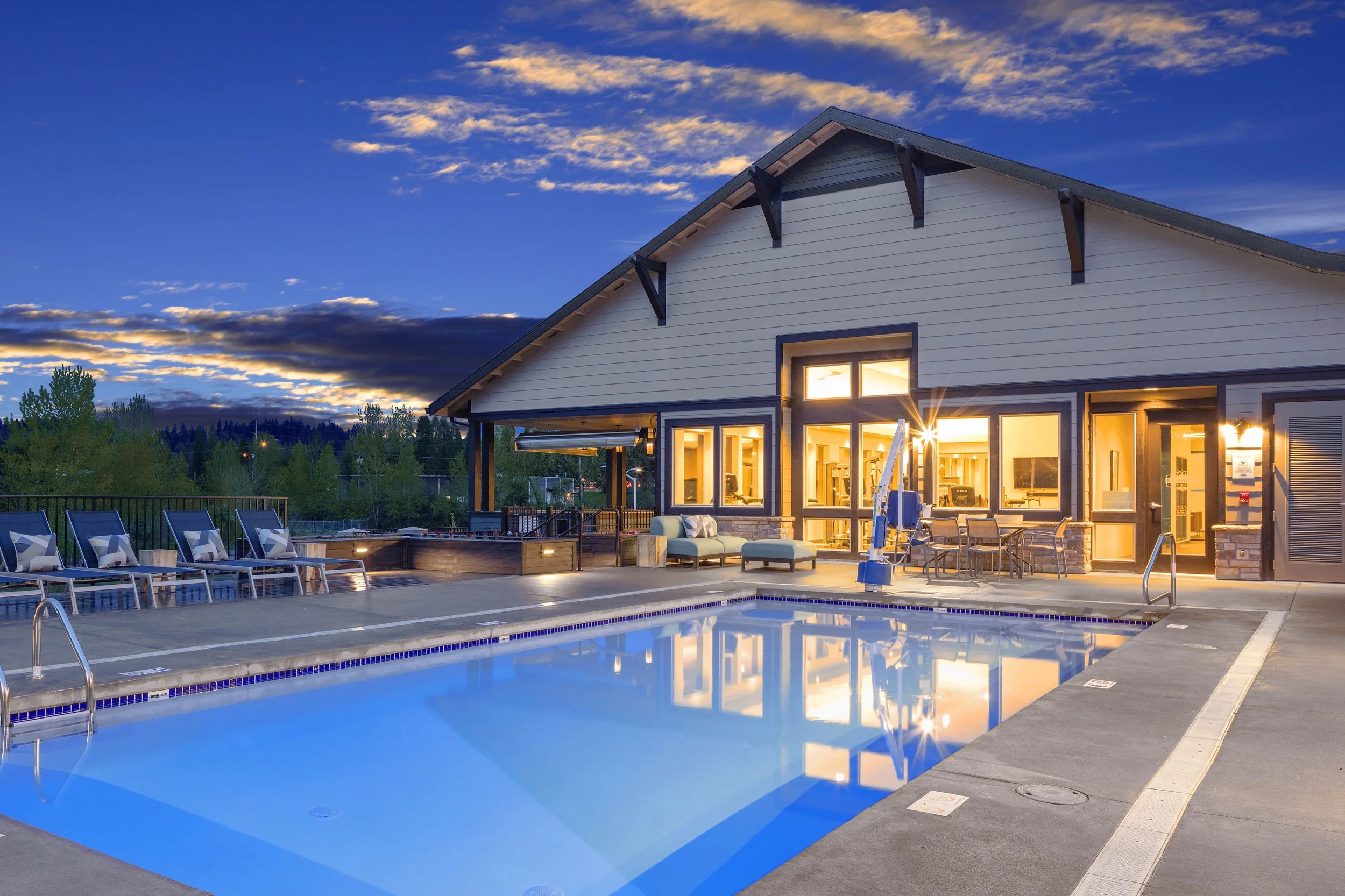 A swimming pool area at dusk with surrounding lounge chairs and a clubhouse with large windows glowing with interior lights.