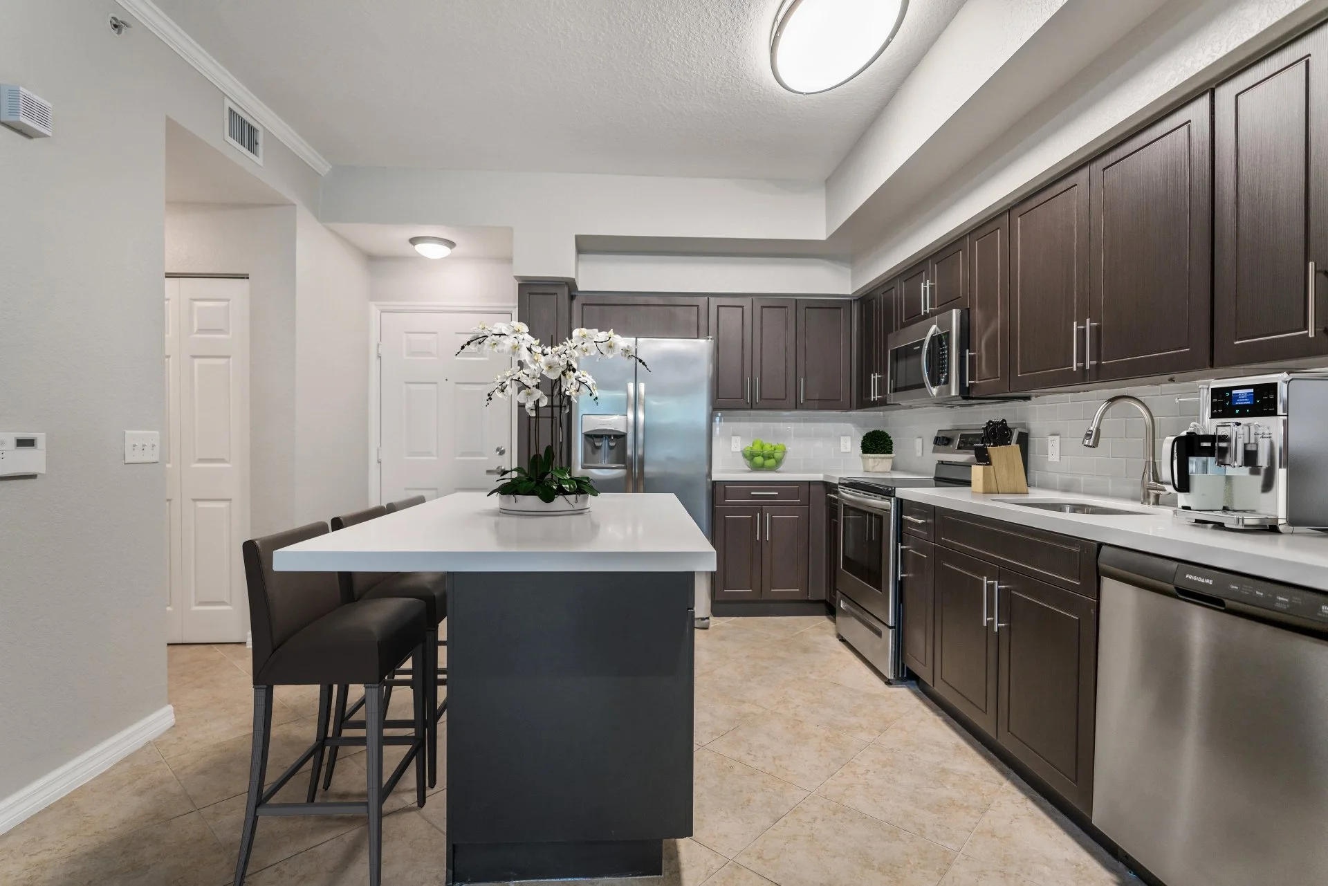 Modern kitchen with dark wooden cabinets, stainless steel appliances including a refrigerator, microwave, oven, dishwasher, and a coffee maker. There is a white island with a vase of white orchids and three black bar stools. The kitchen has beige til