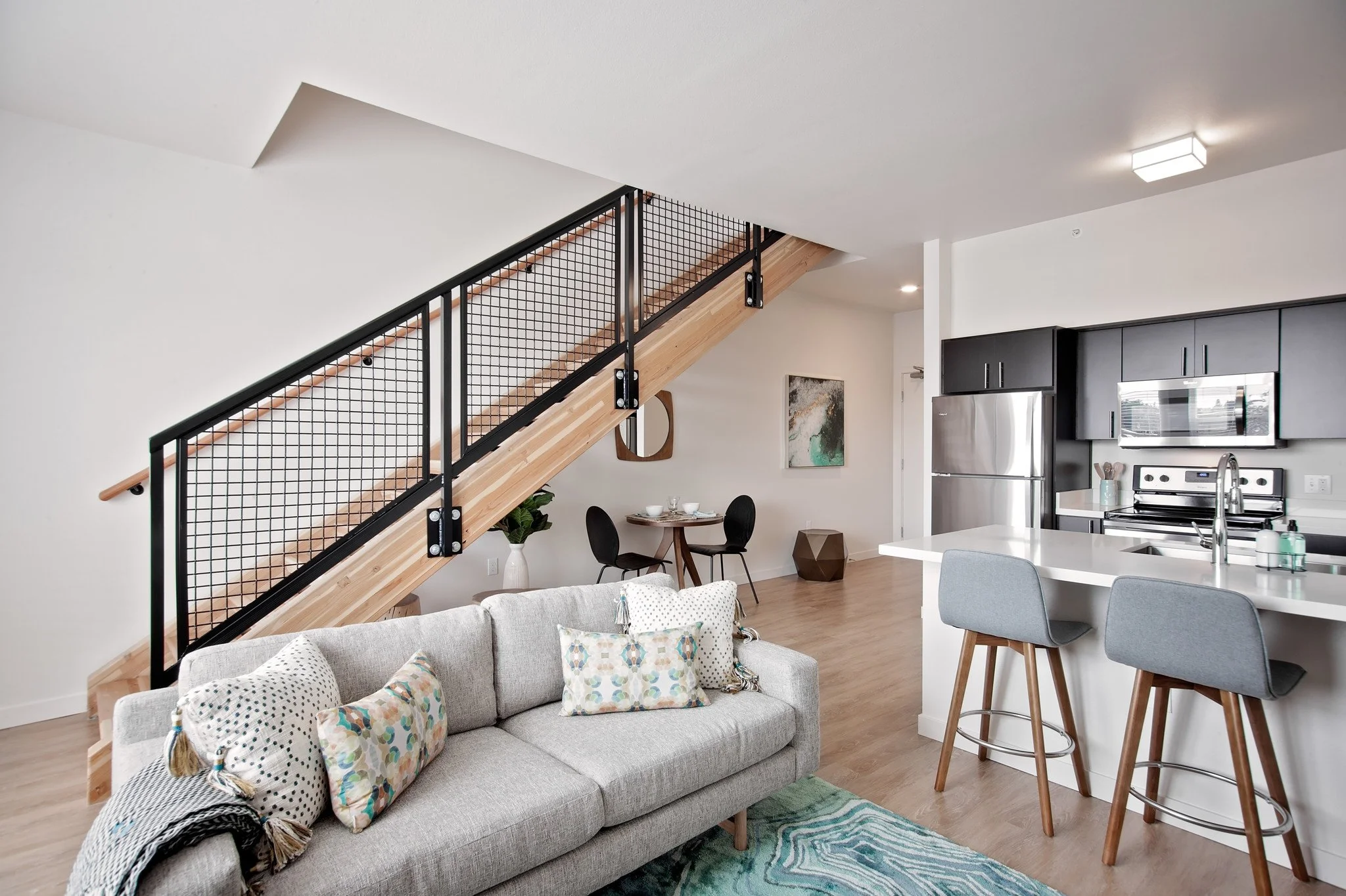 Living room with light gray sofa, decorative pillows, staircase with black metal railing and wooden steps, dining table with black chairs, kitchen with black cabinets, stainless steel appliances, and an island with two gray barstools.