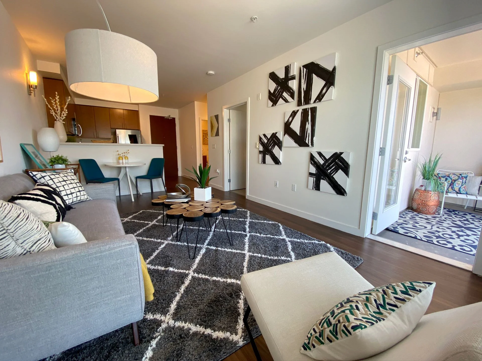Living room with gray sofa, black and white patterned pillows, beige ottoman, black and white geometric rug, modern black-and-white abstract wall art, circular coffee table with wooden segments, potted plant, open area leading to balcony with outdoor