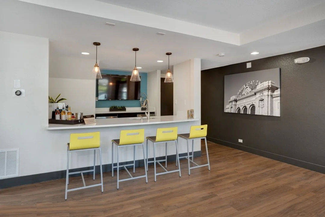 Modern kitchen with four yellow bar stools at a white island, pendant lighting, a mounted TV, and a black and white photograph of a building on a dark gray wall.