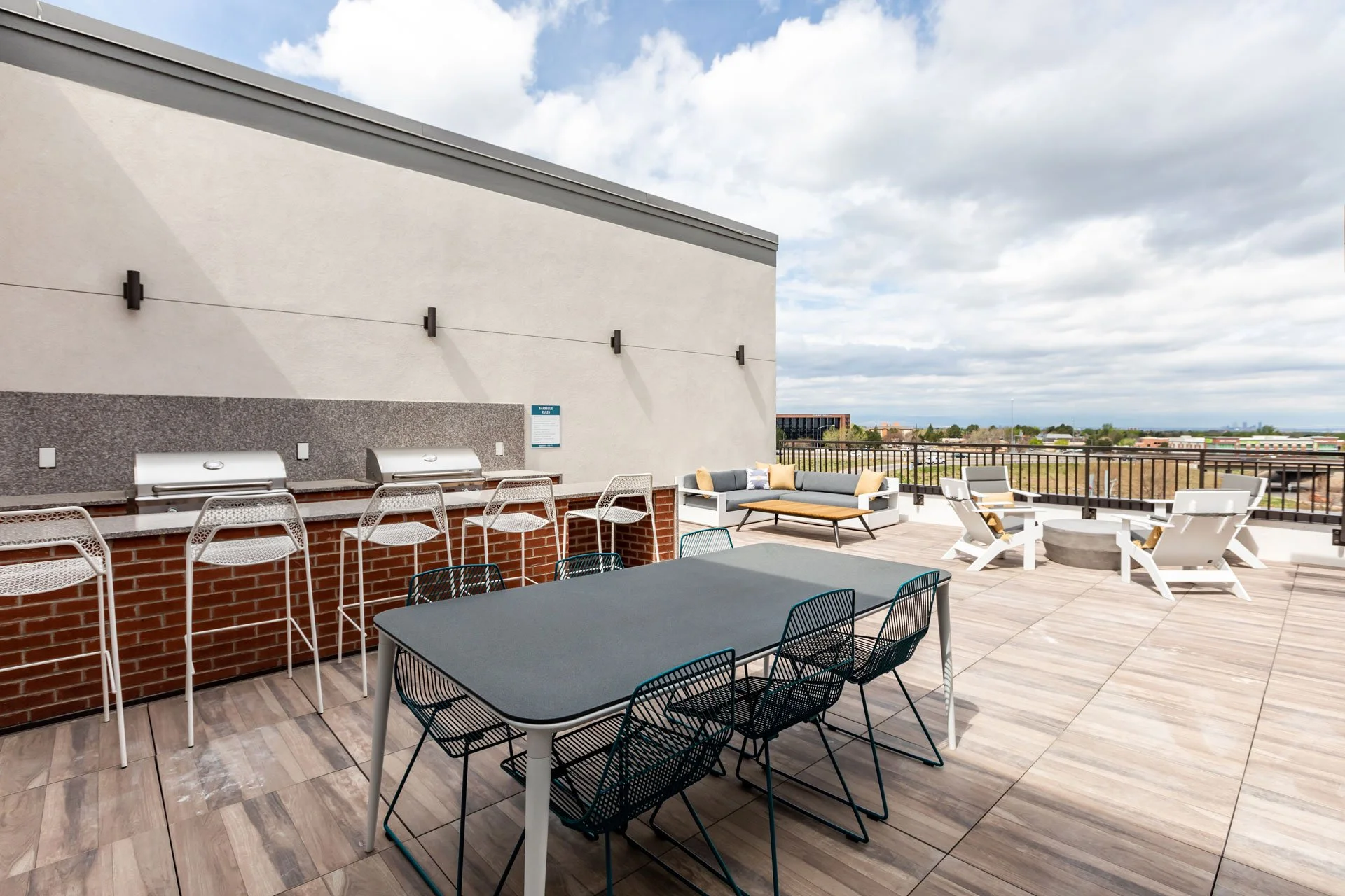 Outdoor rooftop patio with a dining table, black chairs, a seating area with gray and beige sofas, a concrete fire pit, and barbecues, with cityscape view and partly cloudy sky.